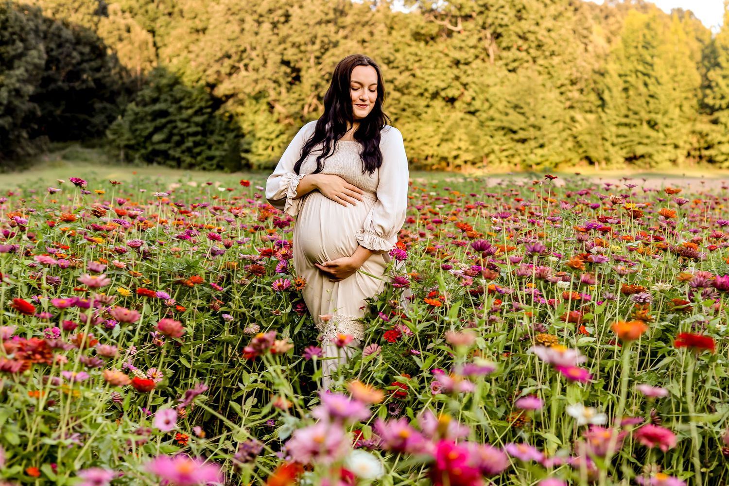 Expectant mother in a white dress poses in a vibrant field of pink wildflowers during golden hour.