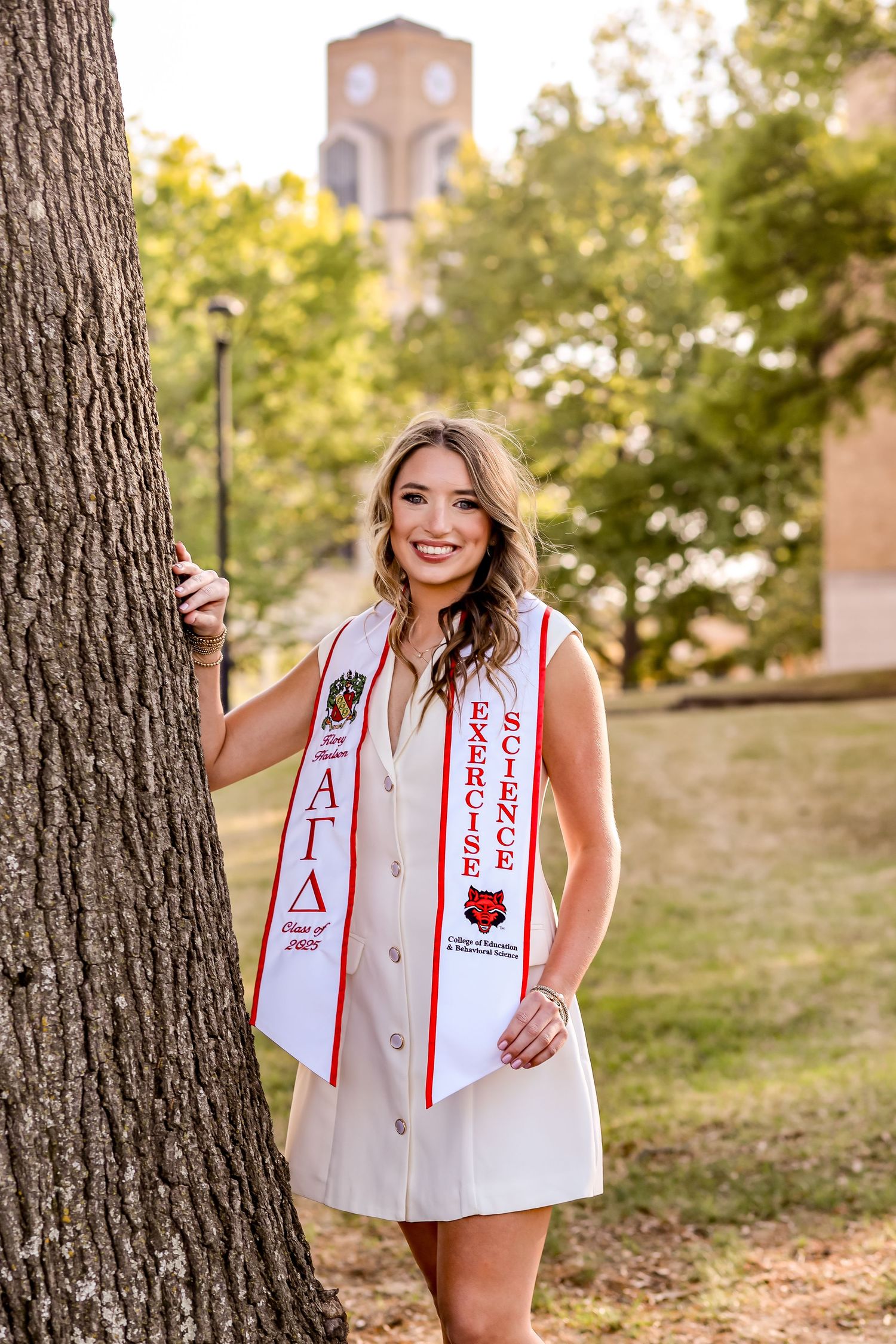 Graduate in white dress and graduation sash poses next to large tree on campus grounds.
