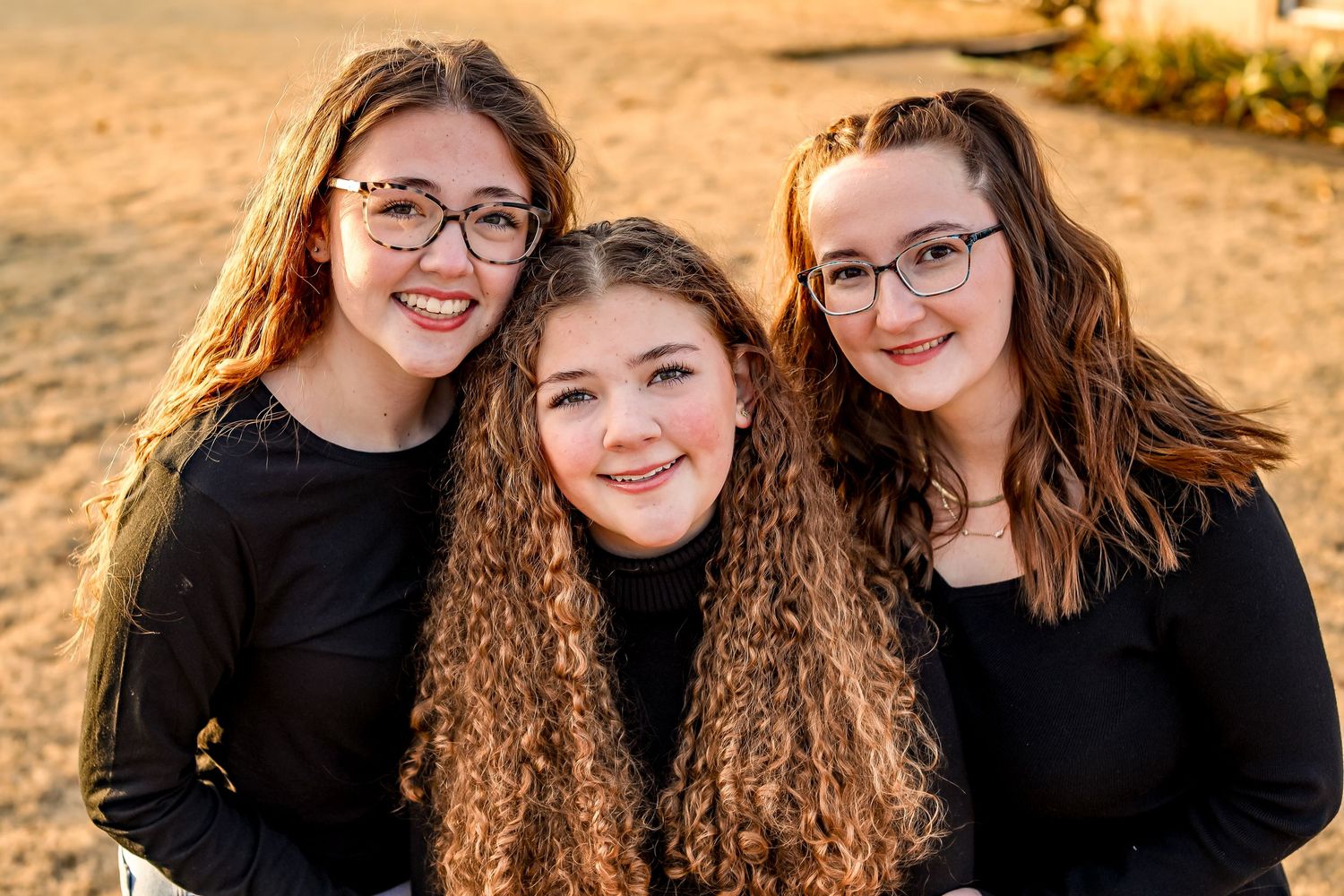 Three young women in black tops pose together at sunset with warm lighting.