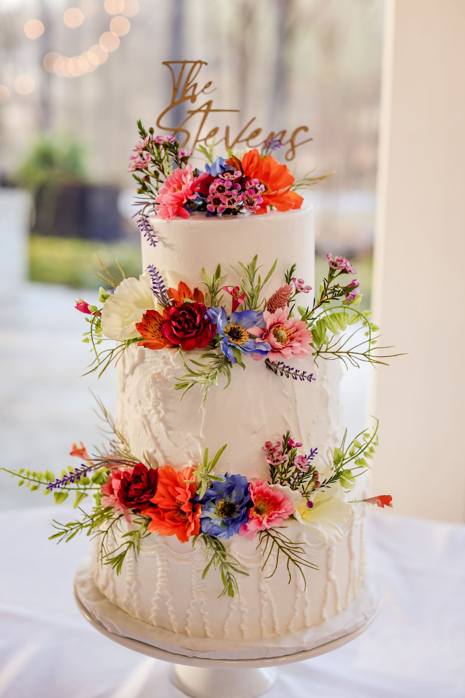Three-tiered white wedding cake decorated with colorful wildflowers and a gold topper.