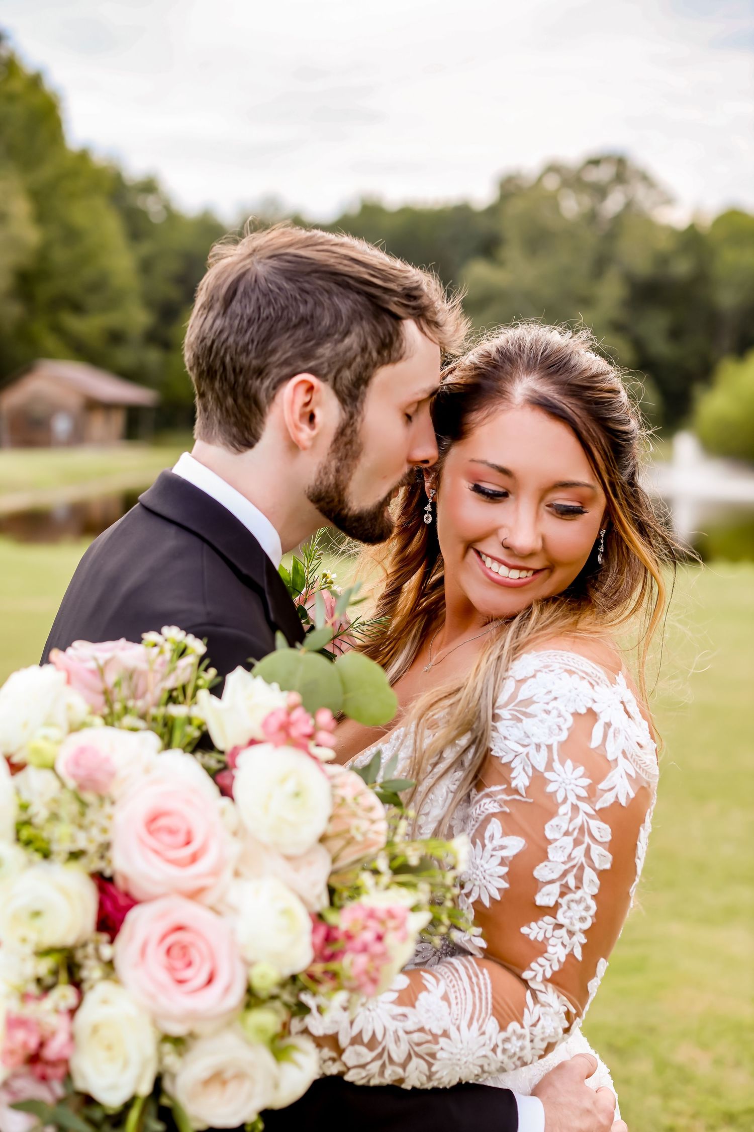 A romantic wedding couple embraces in a sunny field with pink and white rose bouquet.