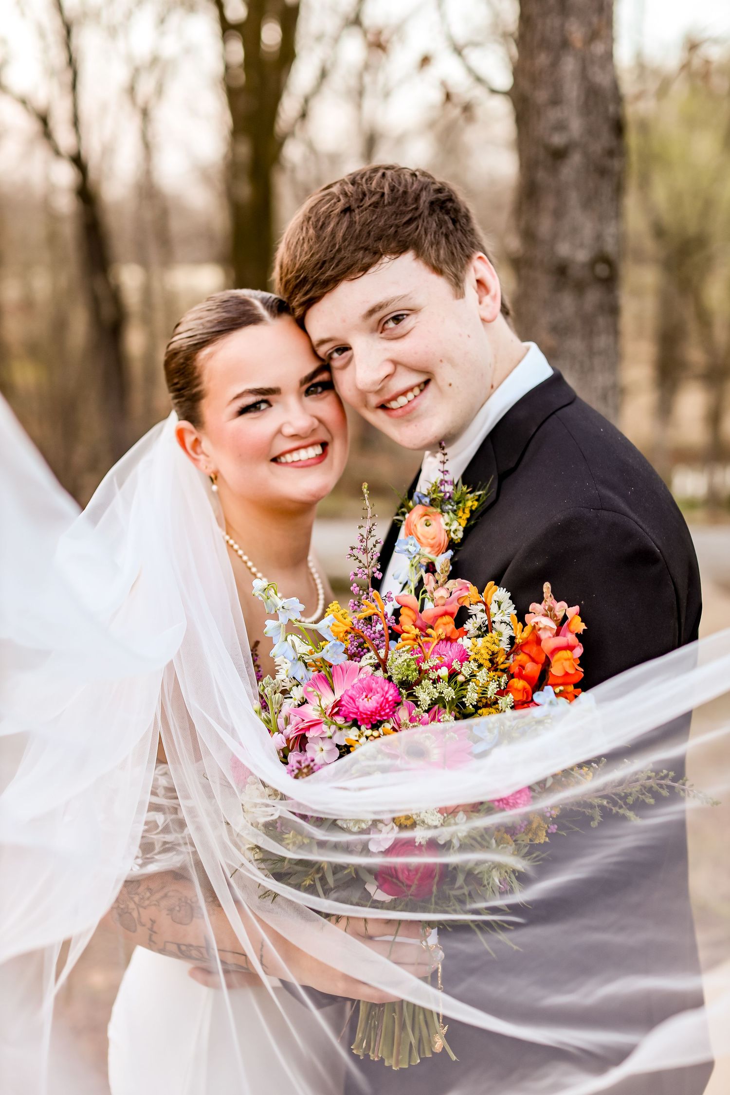 Wedding couple shares romantic moment with colorful bouquet and flowing veil in natural setting.