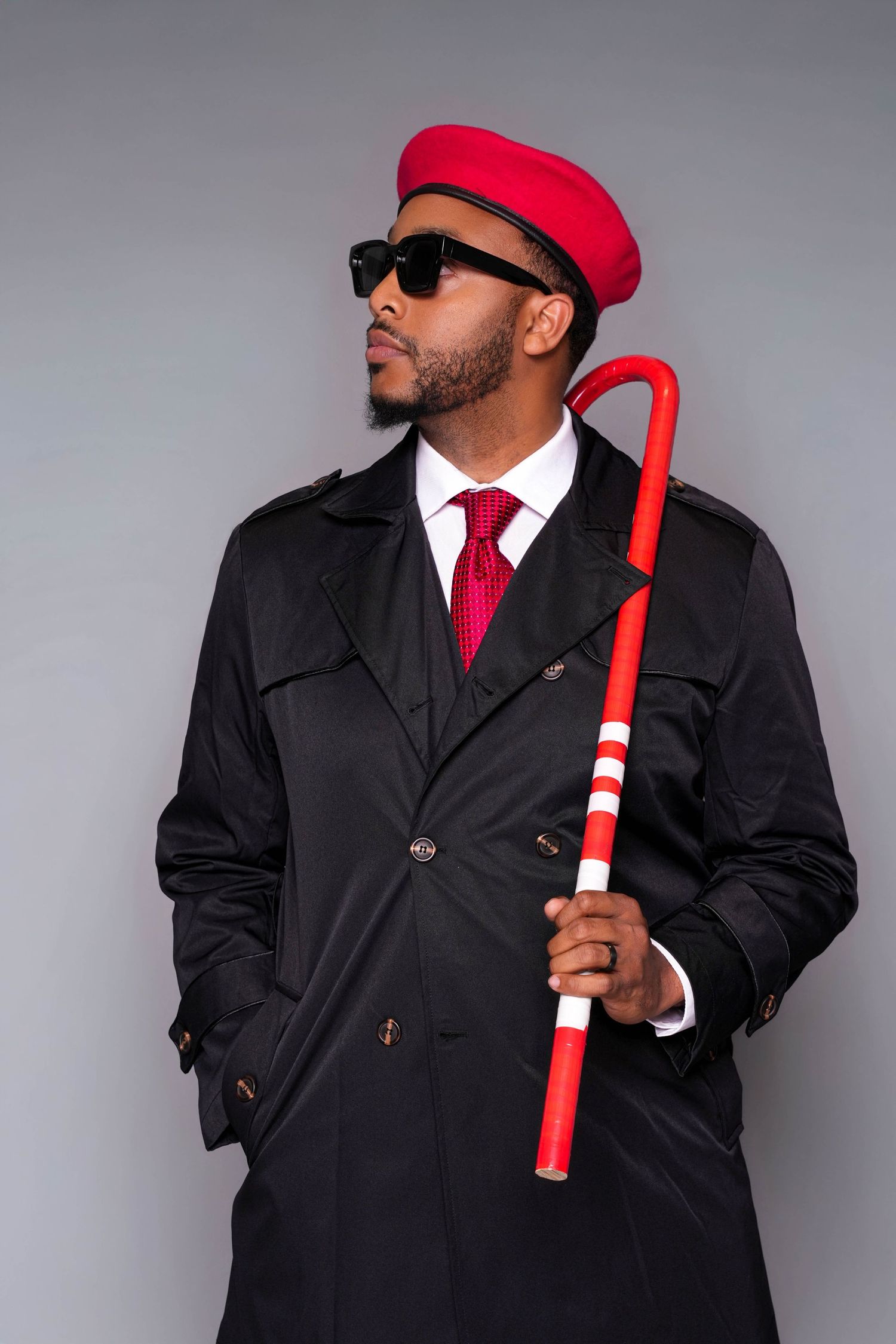 Stylish man in black trench coat, red cap, sunglasses, red tie, holding a red and white cane against gray background.