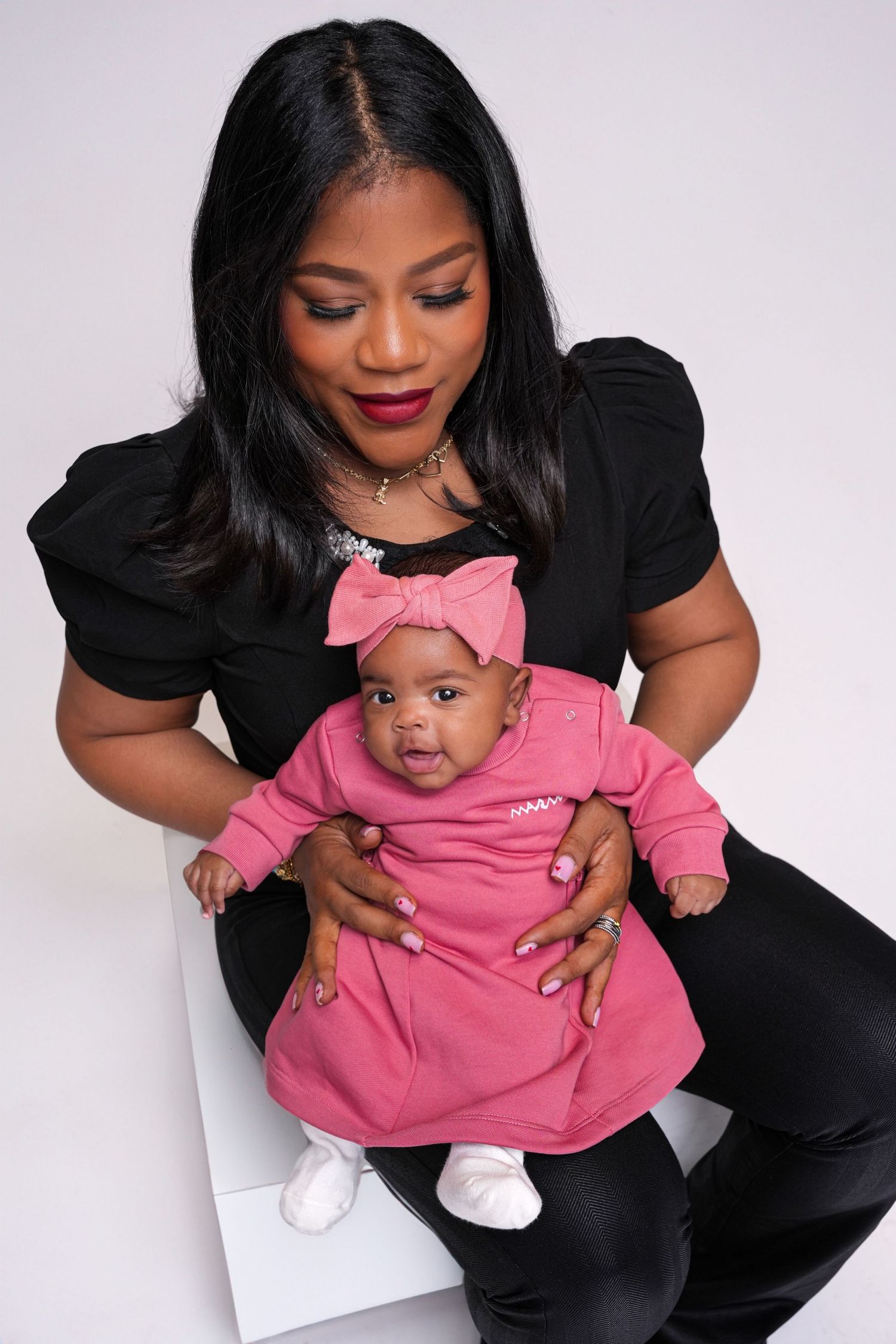 A parent in a black outfit holds a baby wearing a pink dress and bow against a light background.