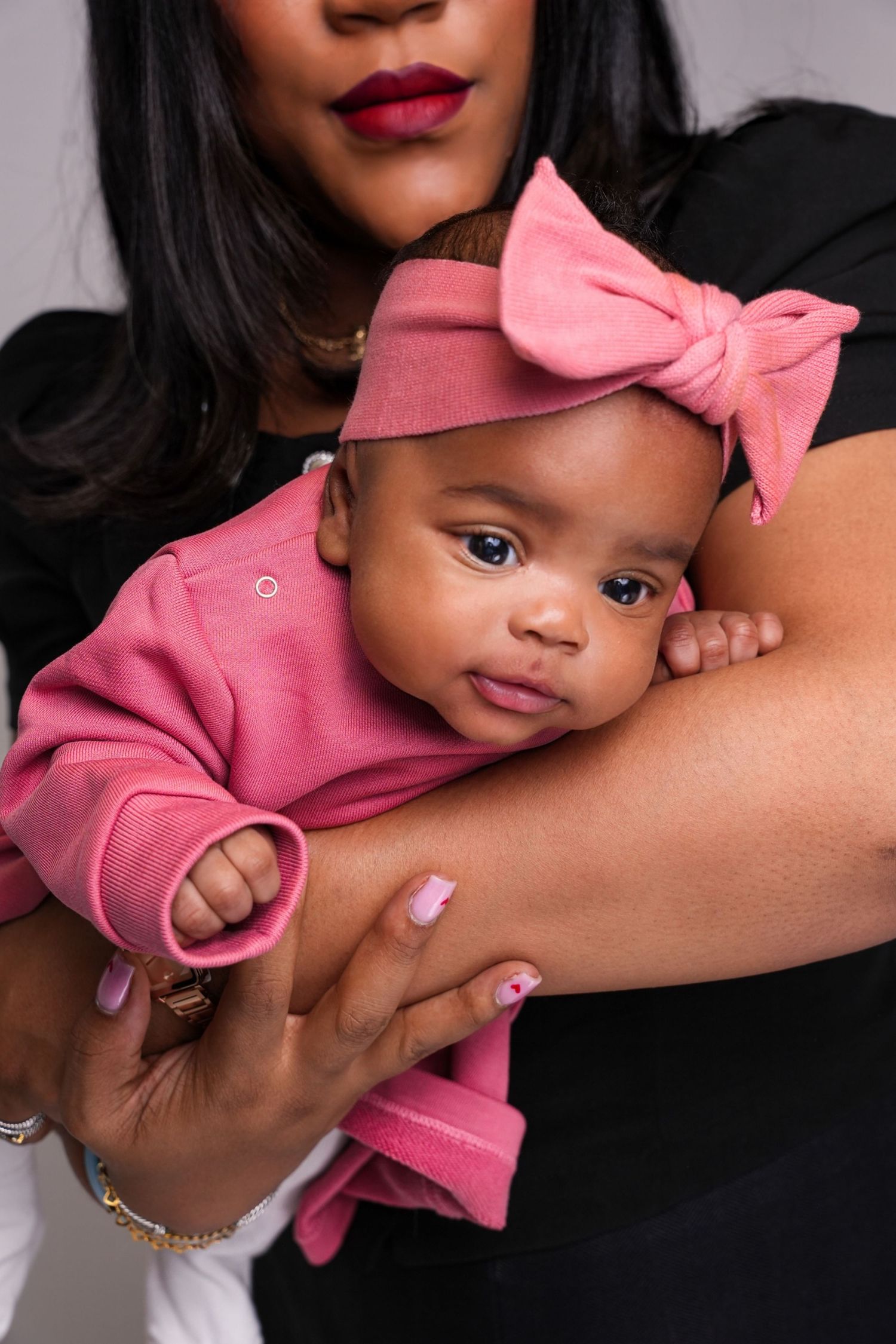 A baby wearing a pink outfit with matching bow headband is held lovingly in someone's arms.
