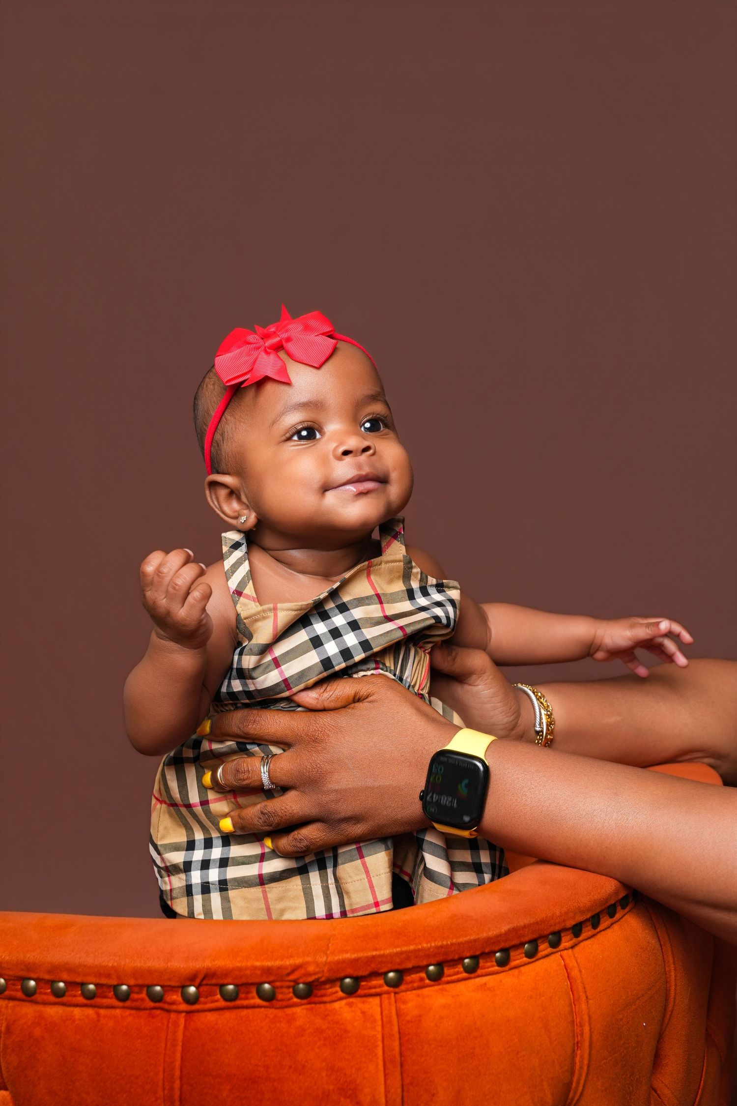 Adorable baby girl in Burberry dress and red bow headband, smiling while being held on an orange velvet ottoman.