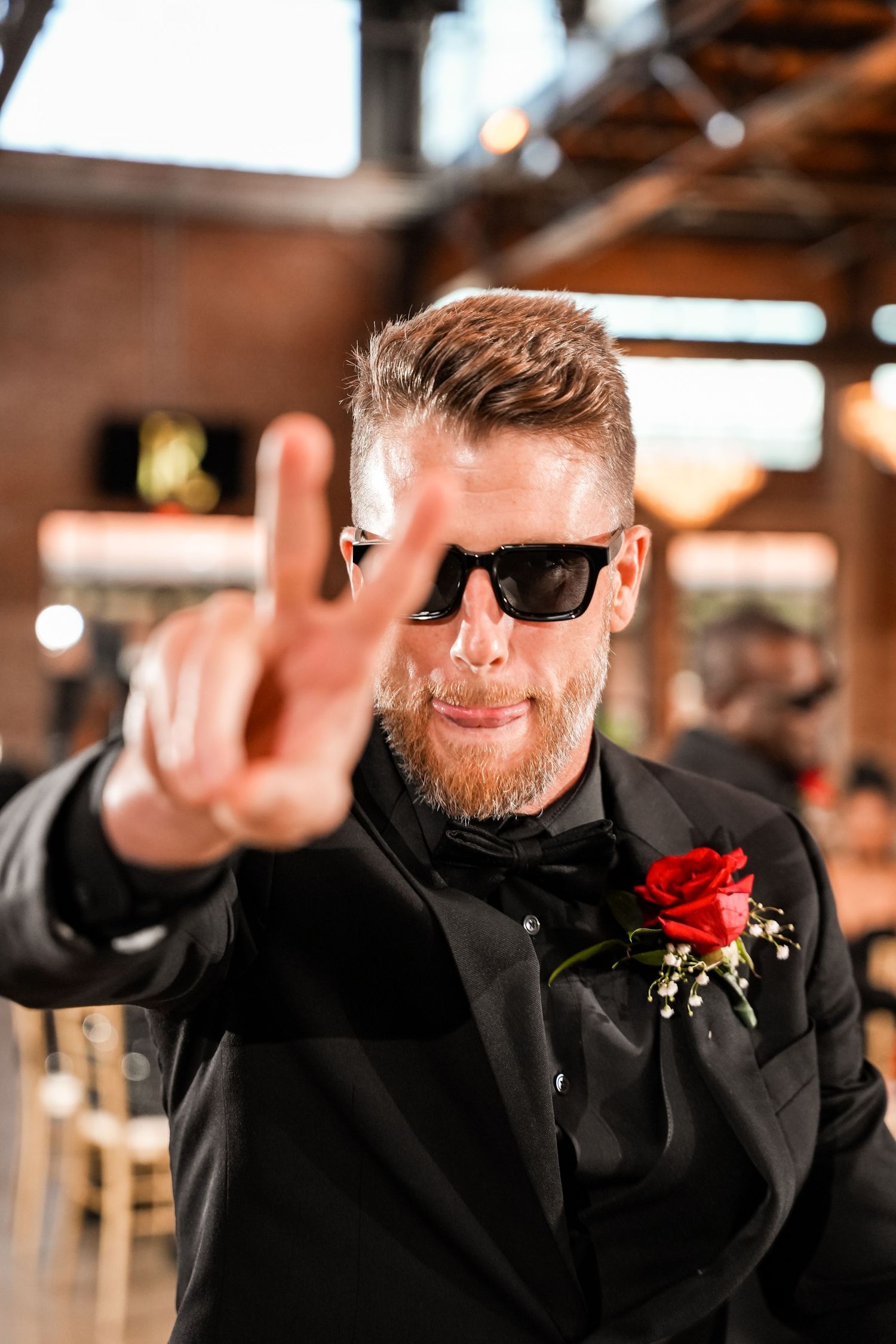 A person in a black suit with sunglasses and red boutonniere makes a peace sign gesture.