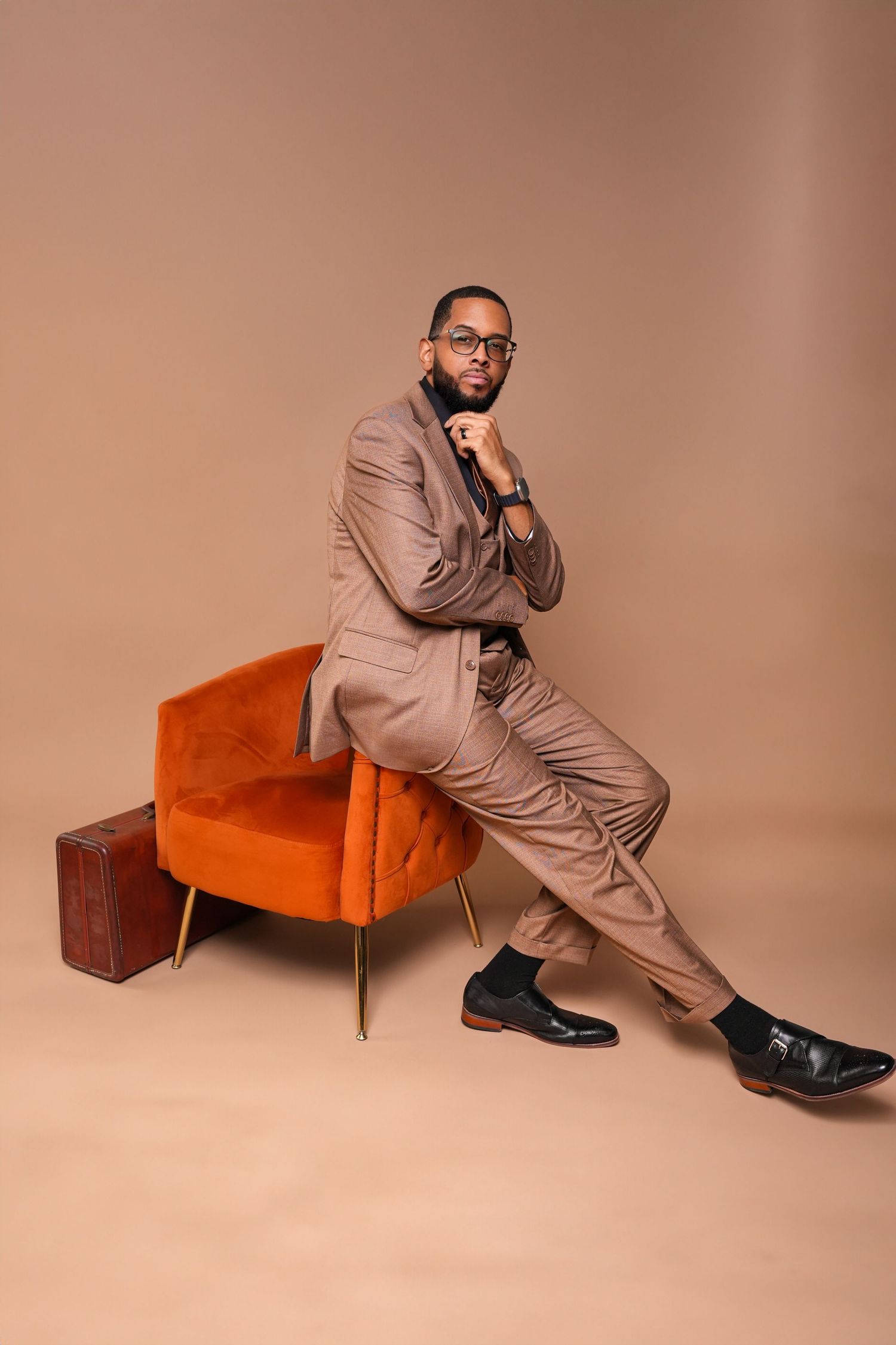 A series of fashion photos showing someone in a beige suit posing with an orange mid-century modern chair against a neutral backdrop.