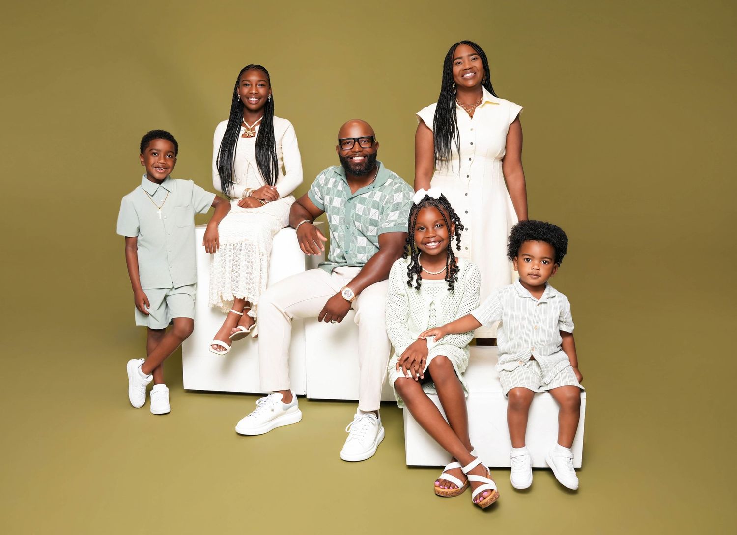 A joyful family of six poses together in coordinated white and green outfits against a warm olive background.