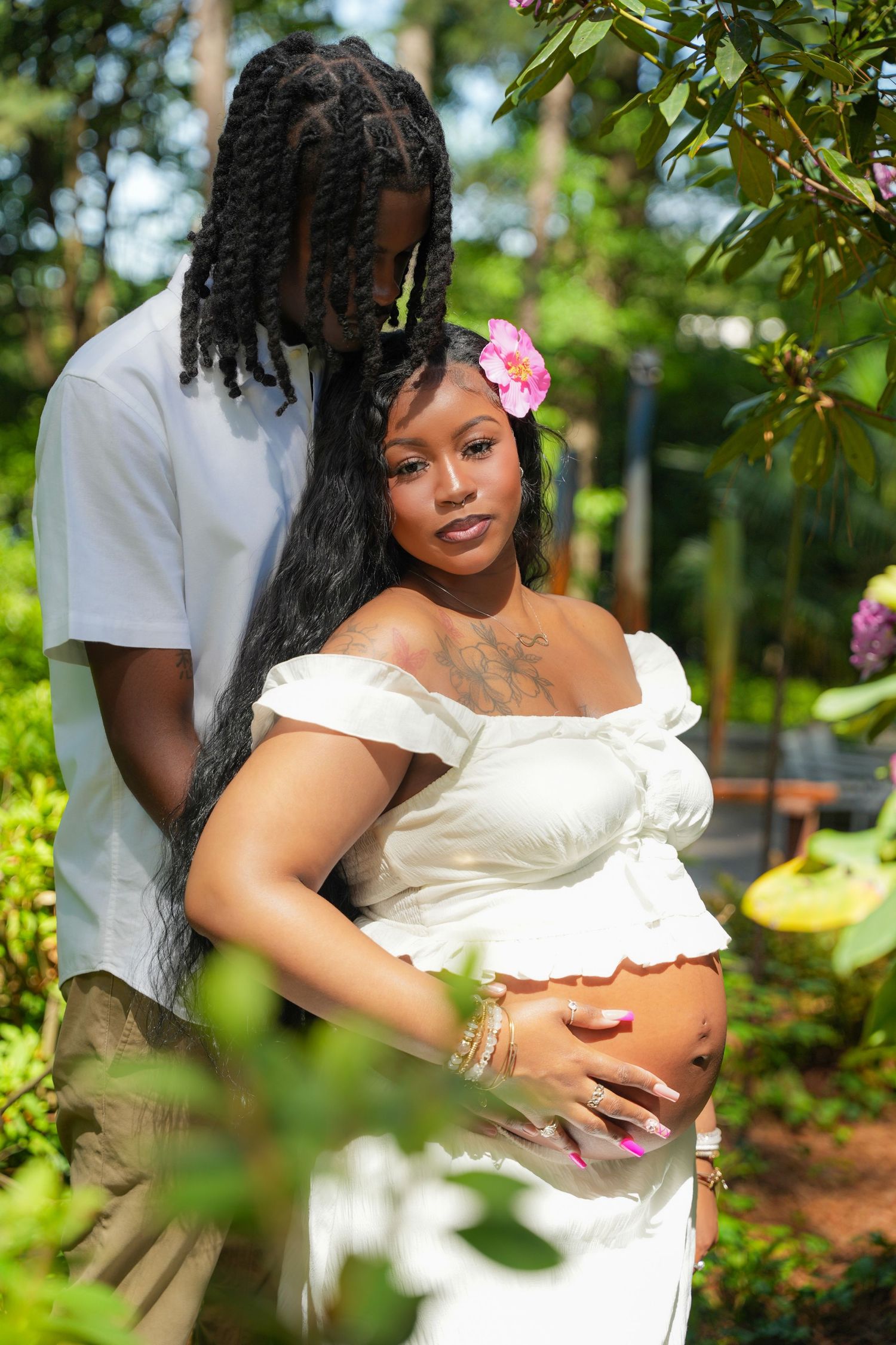 Maternity photo shoot in garden setting with white off-shoulder dress and pink flower accessory.