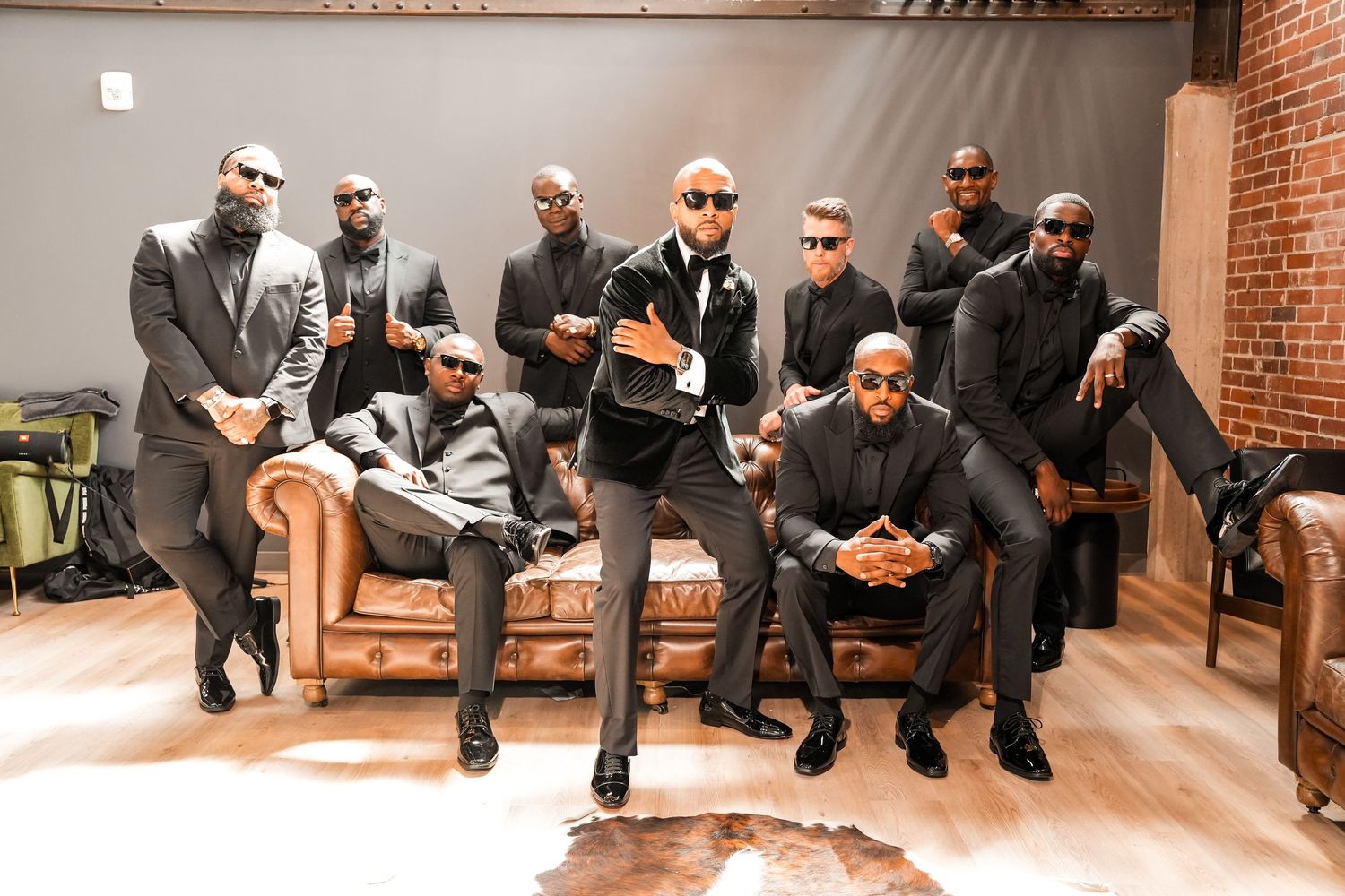 A group in matching black suits and sunglasses pose together on leather furniture.