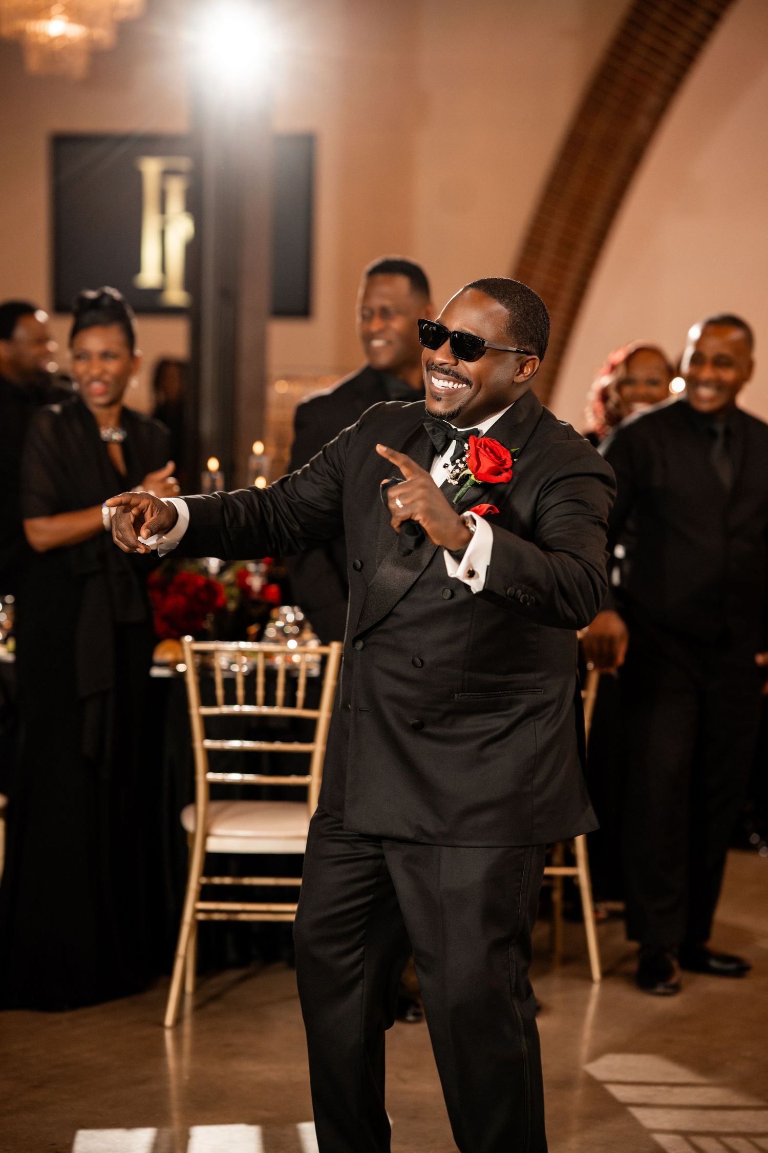 A person in a black tuxedo with red boutonniere dances and sings at an indoor wedding reception.