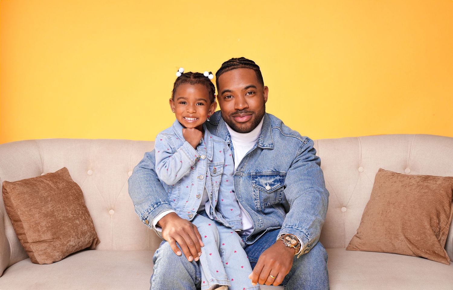 A group of people sitting on a beige couch against a yellow wall wearing casual denim and neutral tones.
