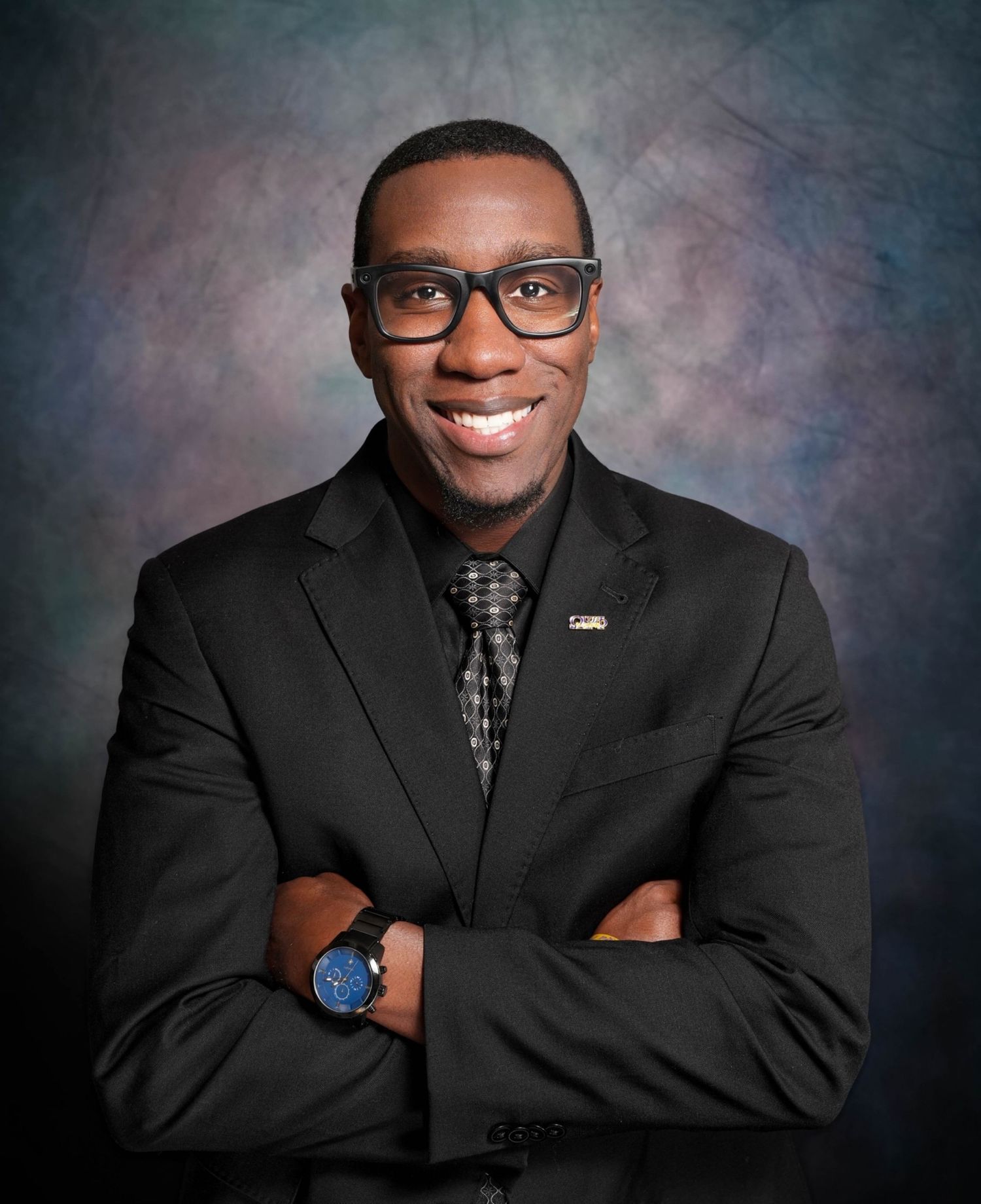 Professional man in black suit with glasses smiling confidently with arms crossed against studio backdrop.