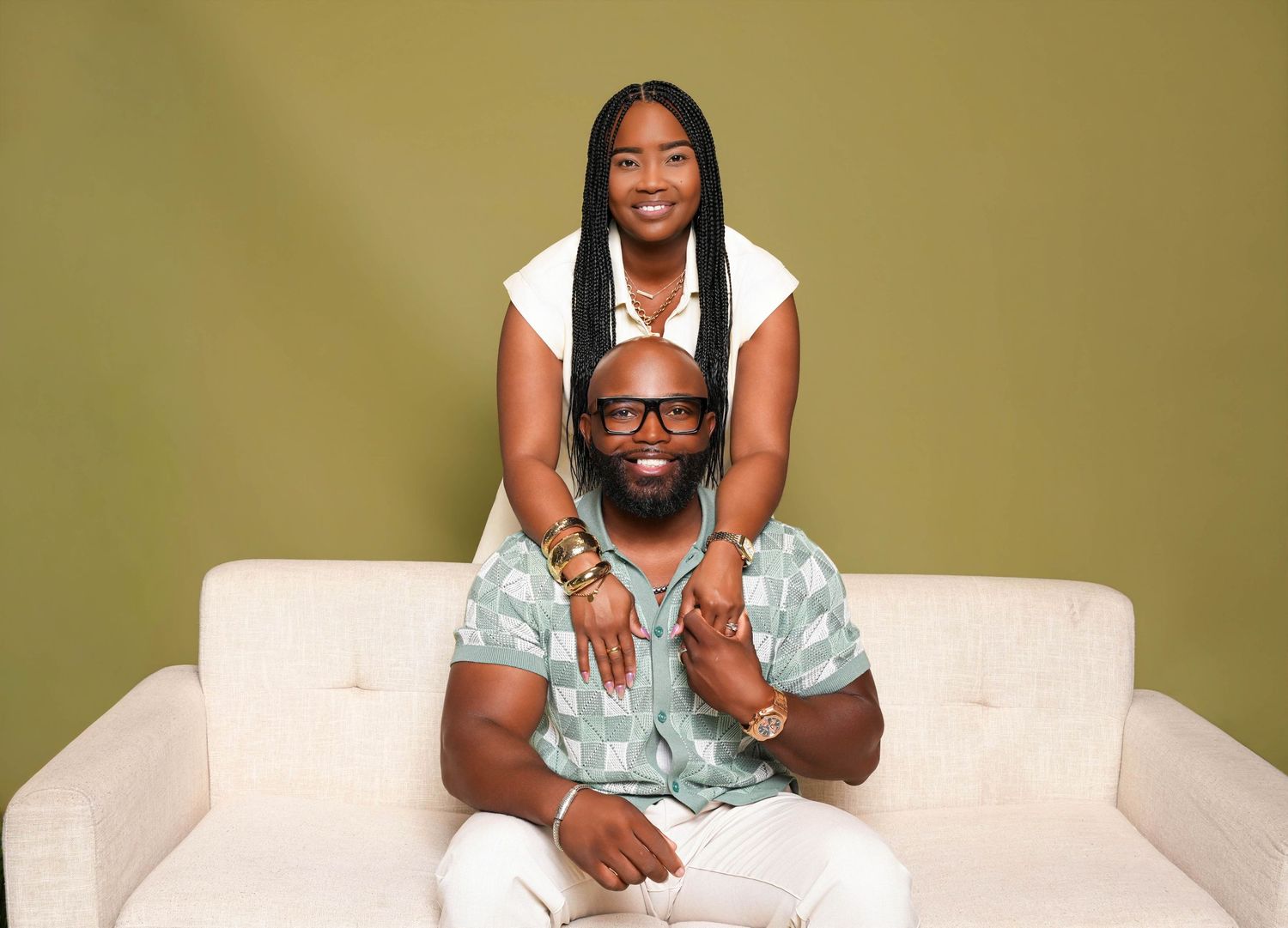 A smiling couple poses together on a white sofa against an olive green background in a professional photo.