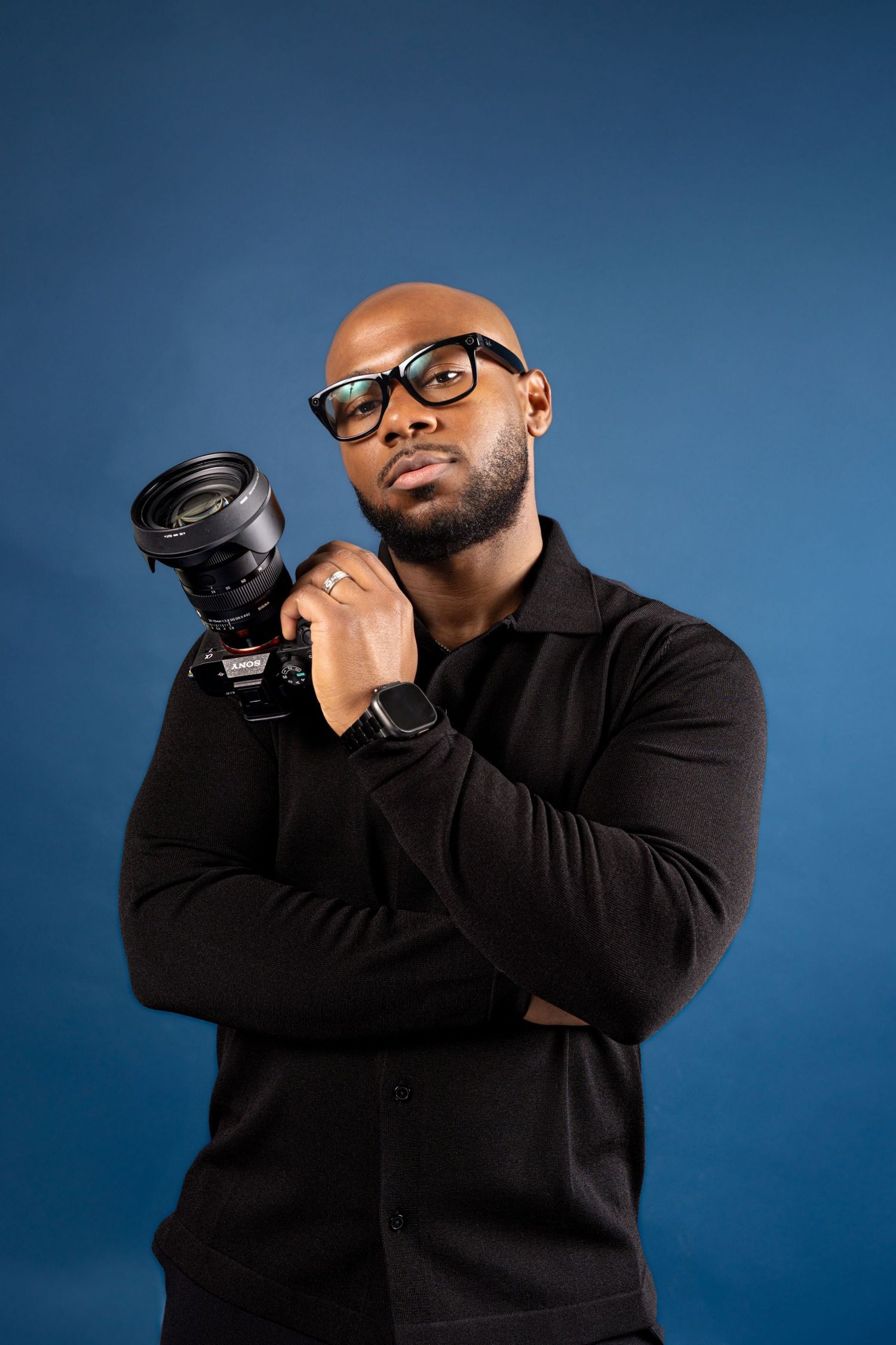 A photographer in black attire holds a professional camera against a blue studio background.