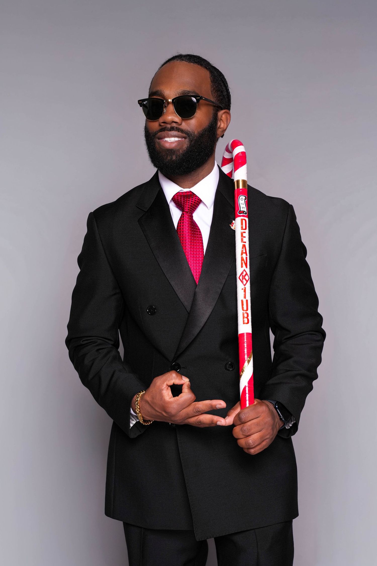 Stylish man in black suit, red tie, sunglasses, holding a decorative candy cane staff, posing confidently.