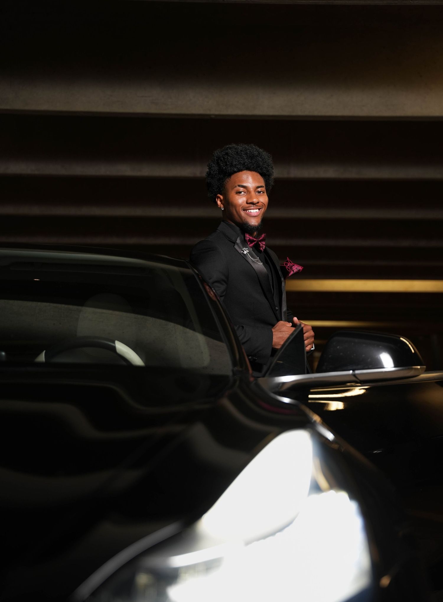 Young man in black tuxedo with red bow tie smiling beside a black car in a parking garage.