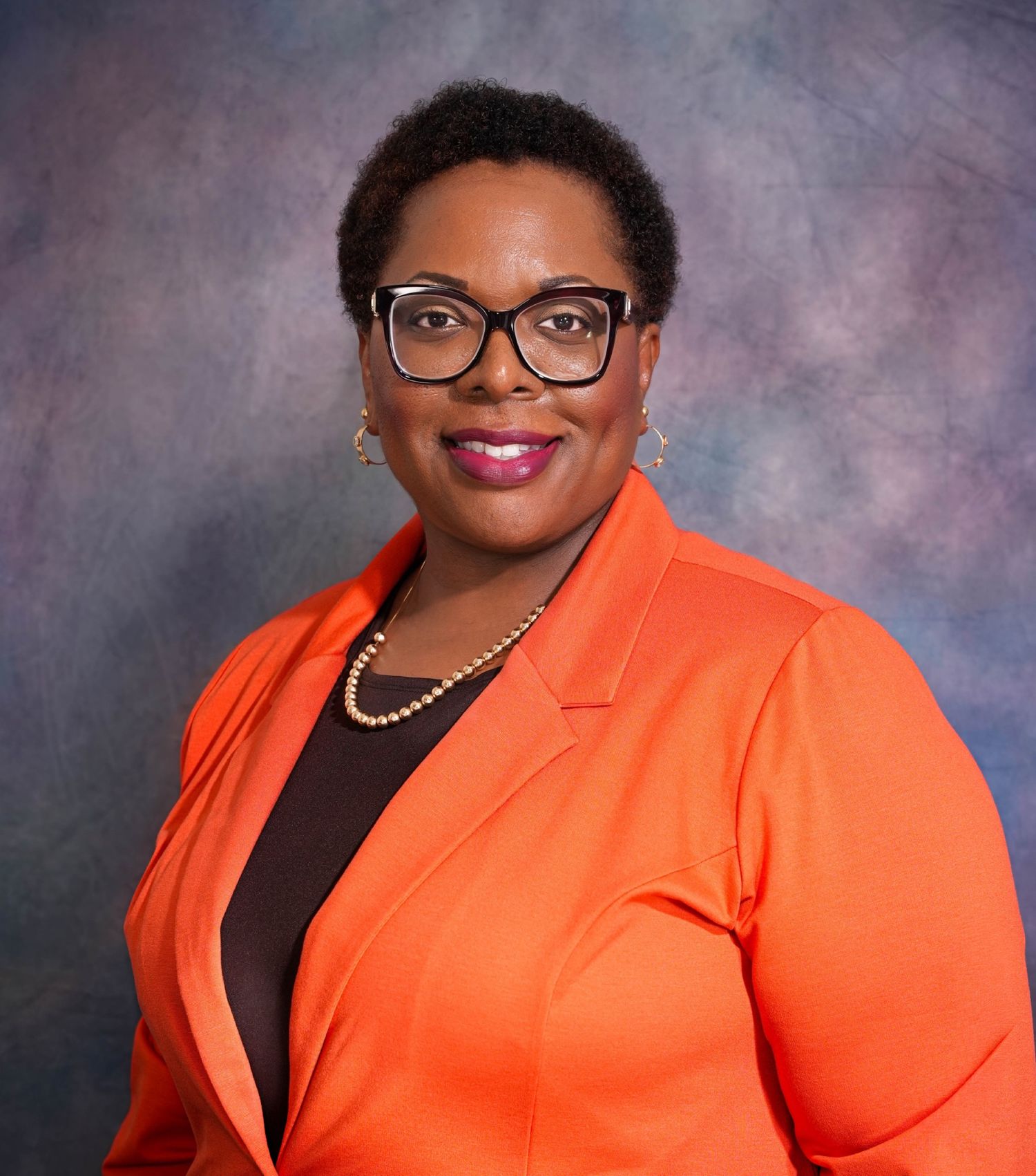 Professional headshot of a woman in an orange blazer and black glasses, smiling confidently against a studio background.