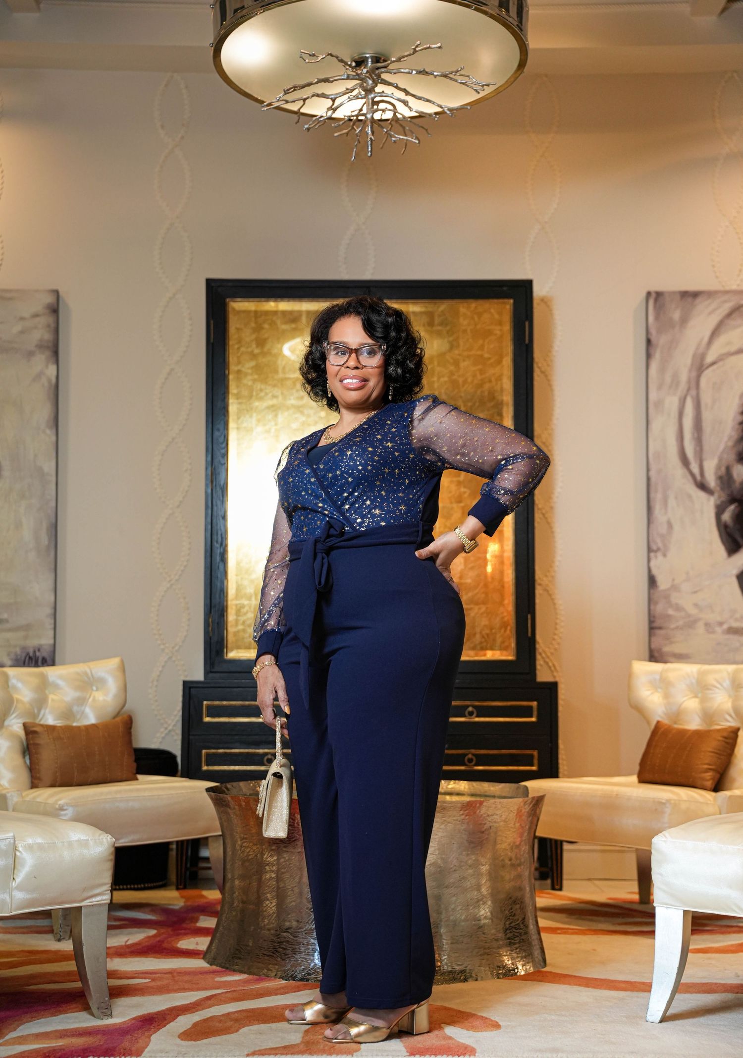 A plus-size model in a navy sequined evening gown poses elegantly in a luxurious hotel suite setting.