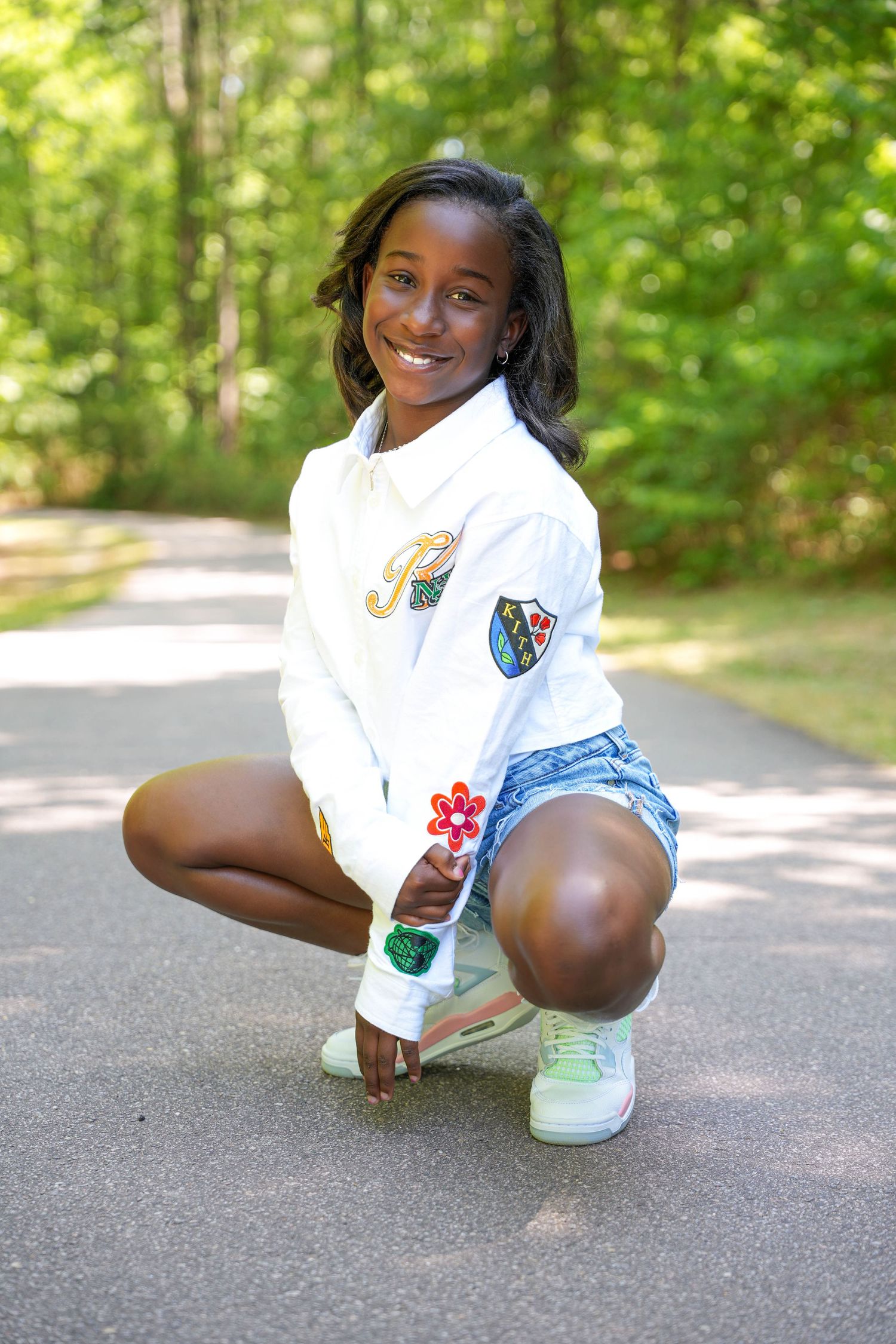 Smiling young girl crouching on a path in a white jacket, denim shorts, and green sneakers, holding a flower lollipop.