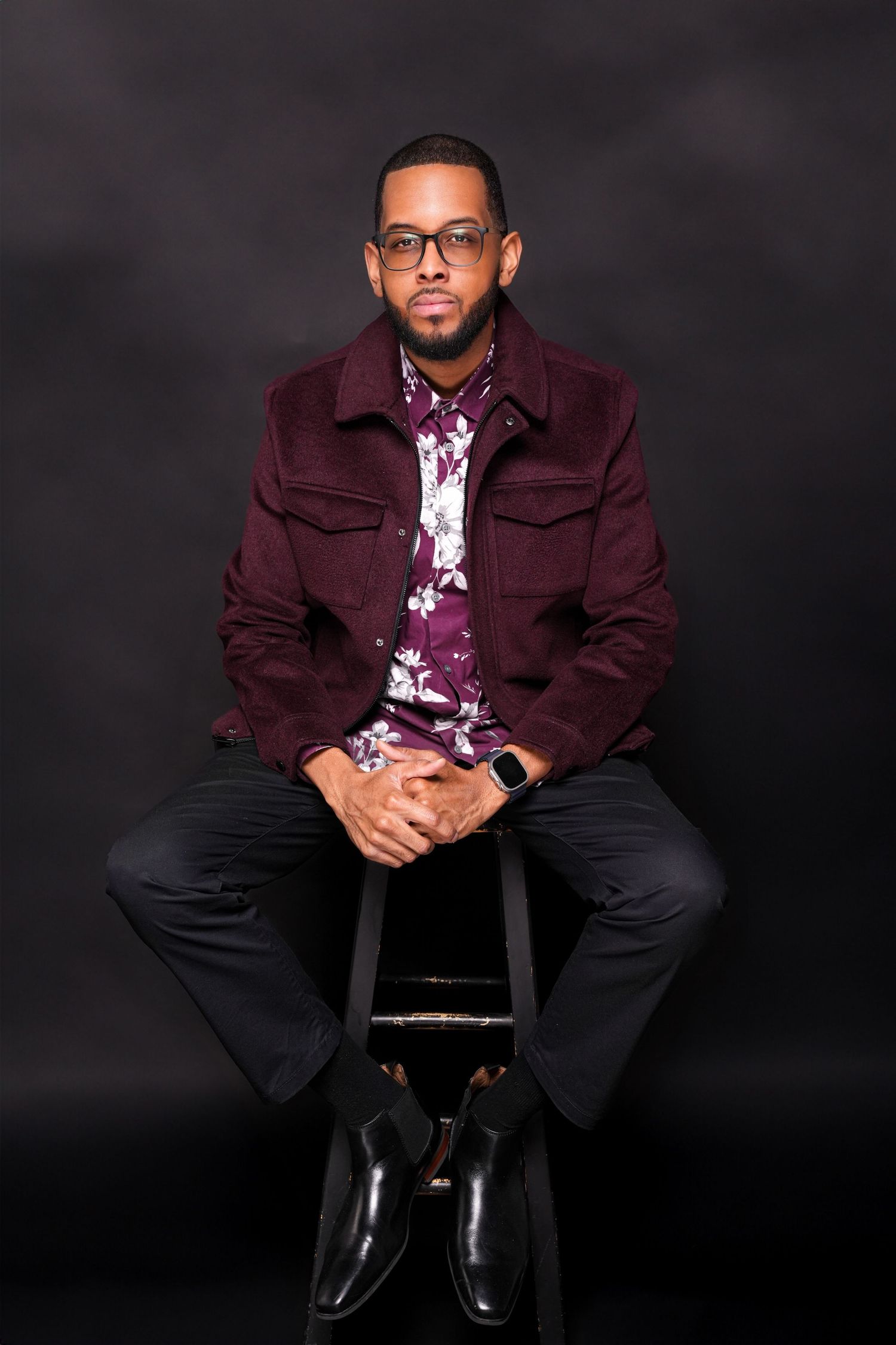 A person in a burgundy jacket and dark pants sits on a stool against a black background in a professional photo shoot.