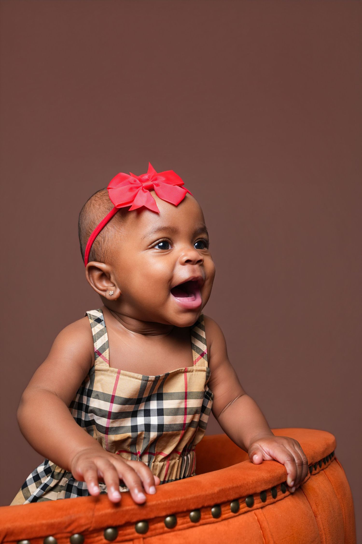 Adorable baby girl in plaid dress and red bow headband laughing joyfully while sitting in an orange chair.