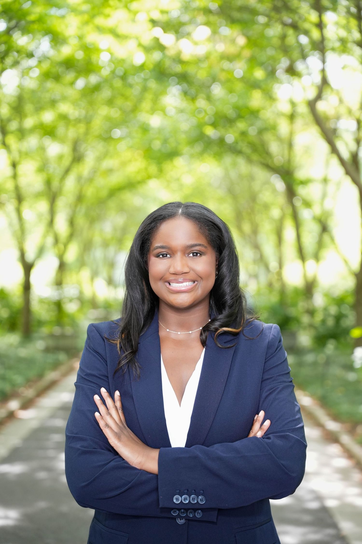 Professional woman in navy blazer smiling with arms crossed in a tree-lined park pathway.
