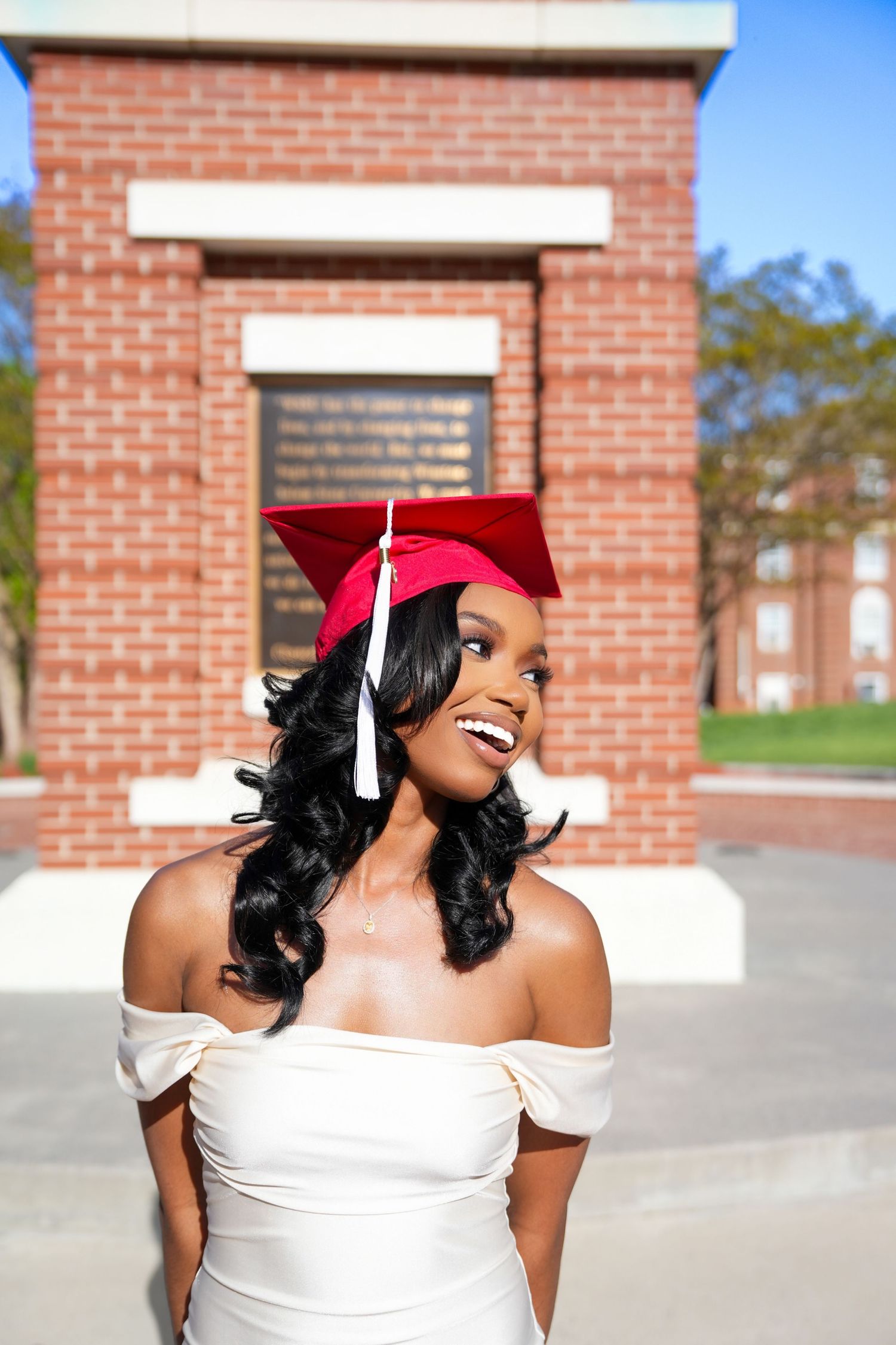 Graduate in white dress and red cap poses joyfully outside brick building.