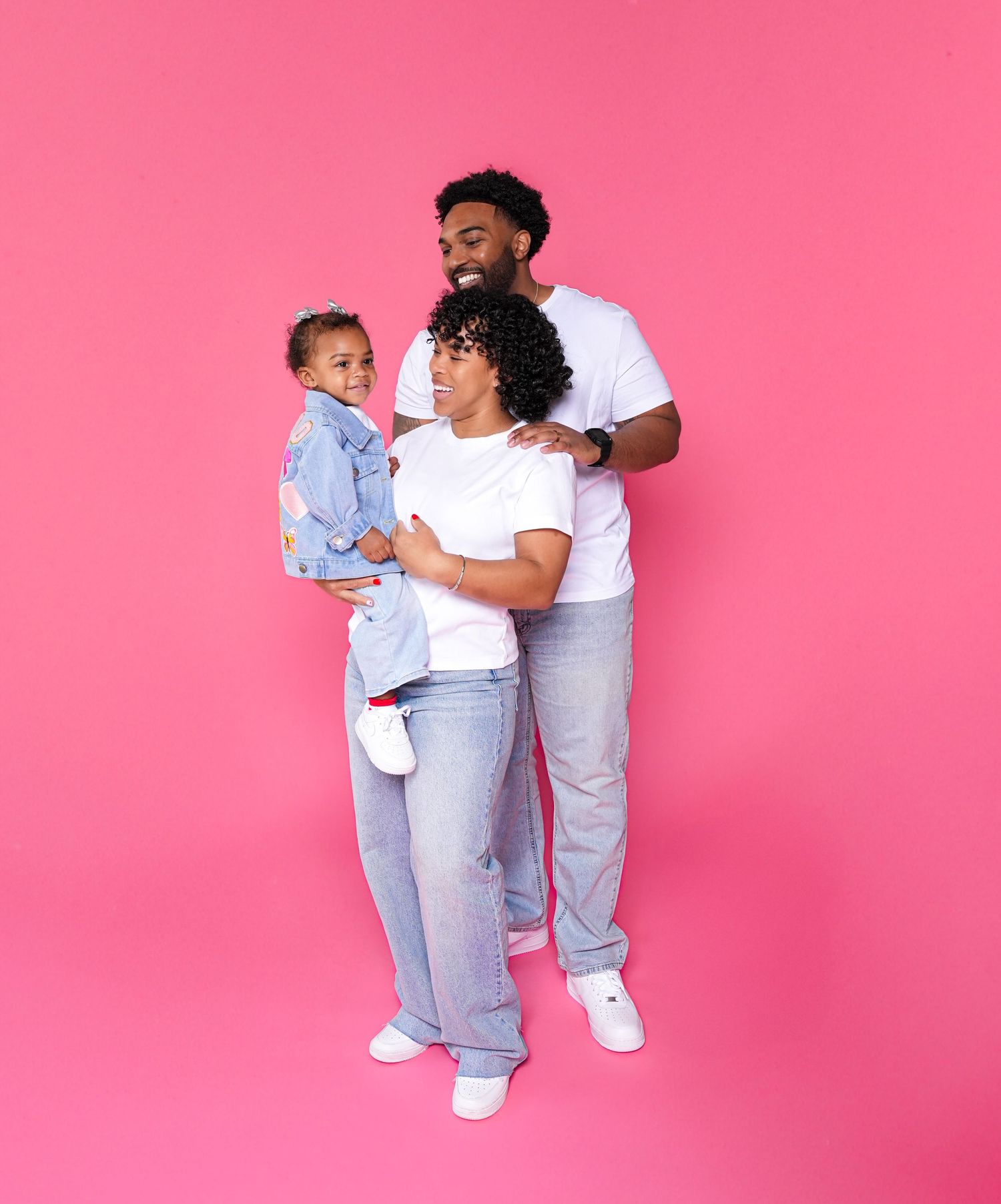 A family wearing matching white shirts and light denim jeans poses together against a bright pink background.
