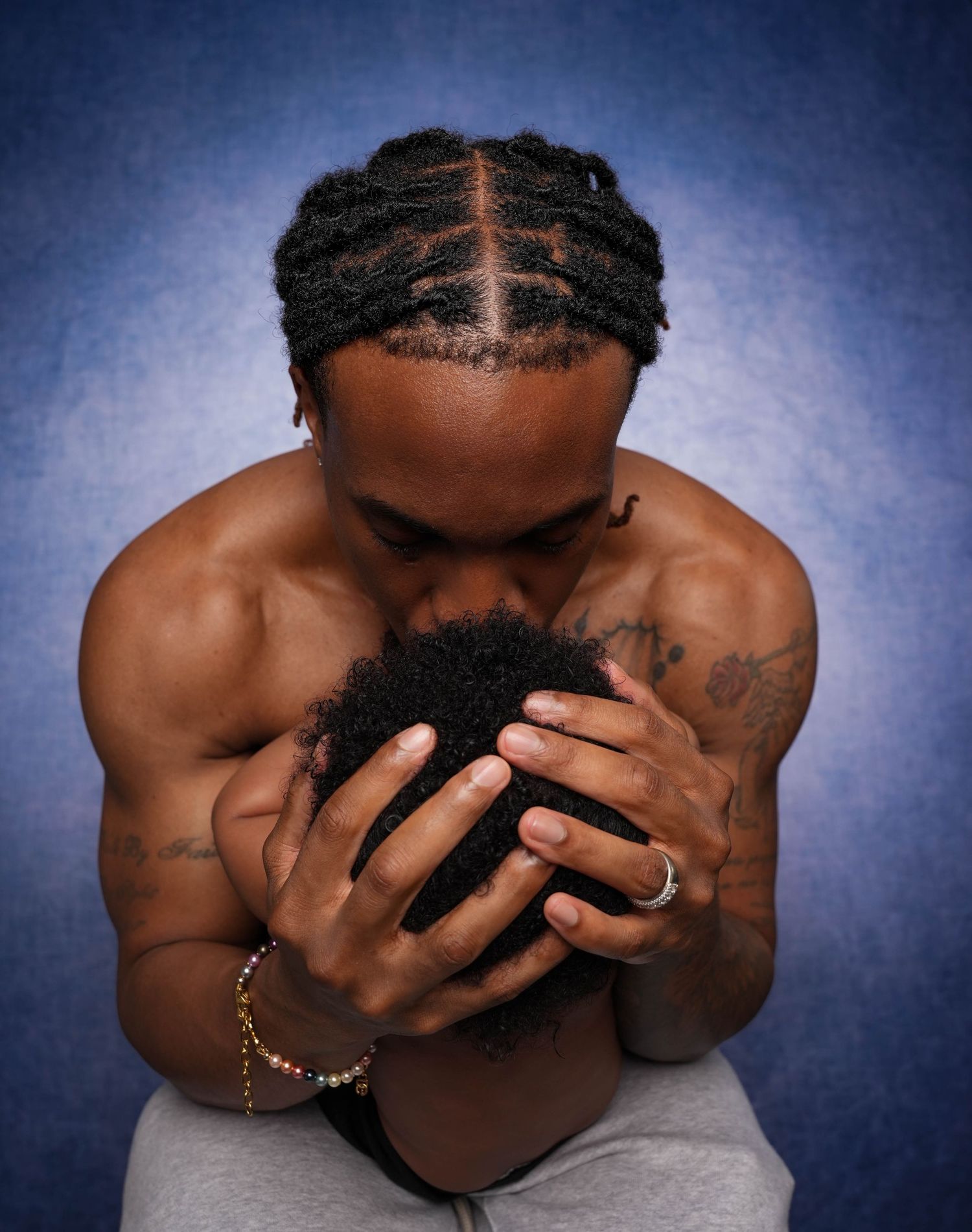 Muscular shirtless man with braided hair holds a child tenderly against his chest in a studio portrait.