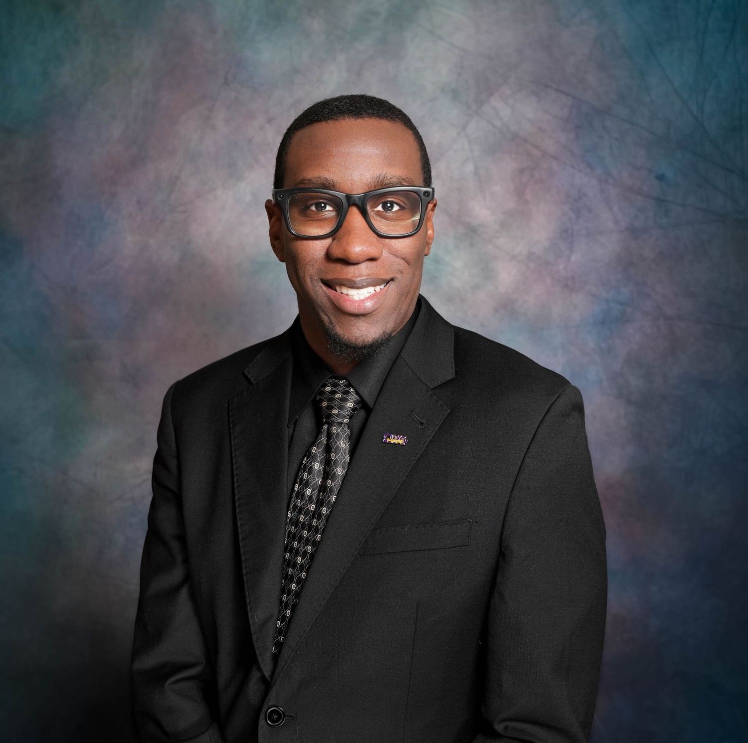 Professional headshot of a young man in a black suit and patterned tie, smiling confidently against a studio backdrop.