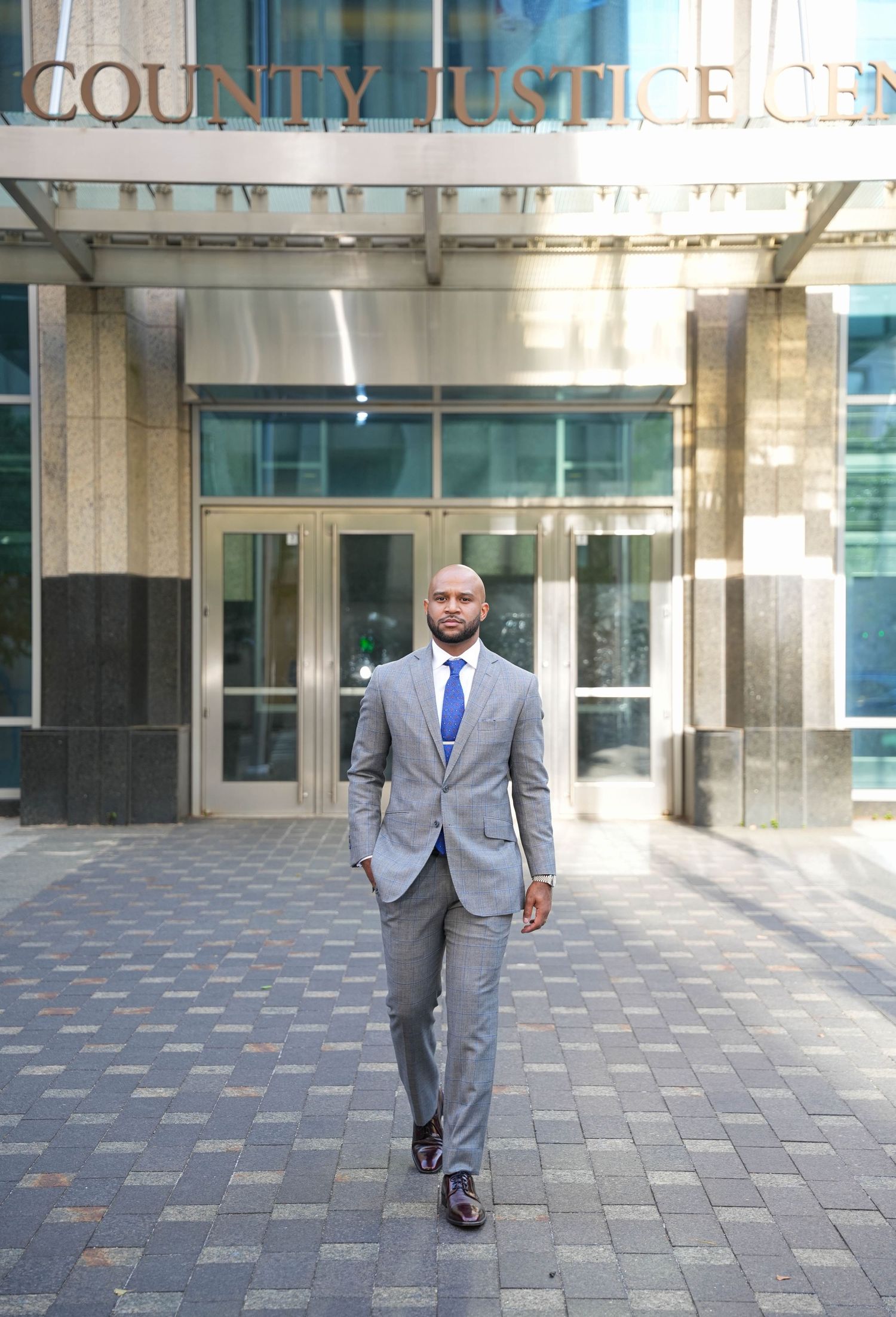 A professional man in a gray suit and blue tie walks confidently outside a County Justice Center building.