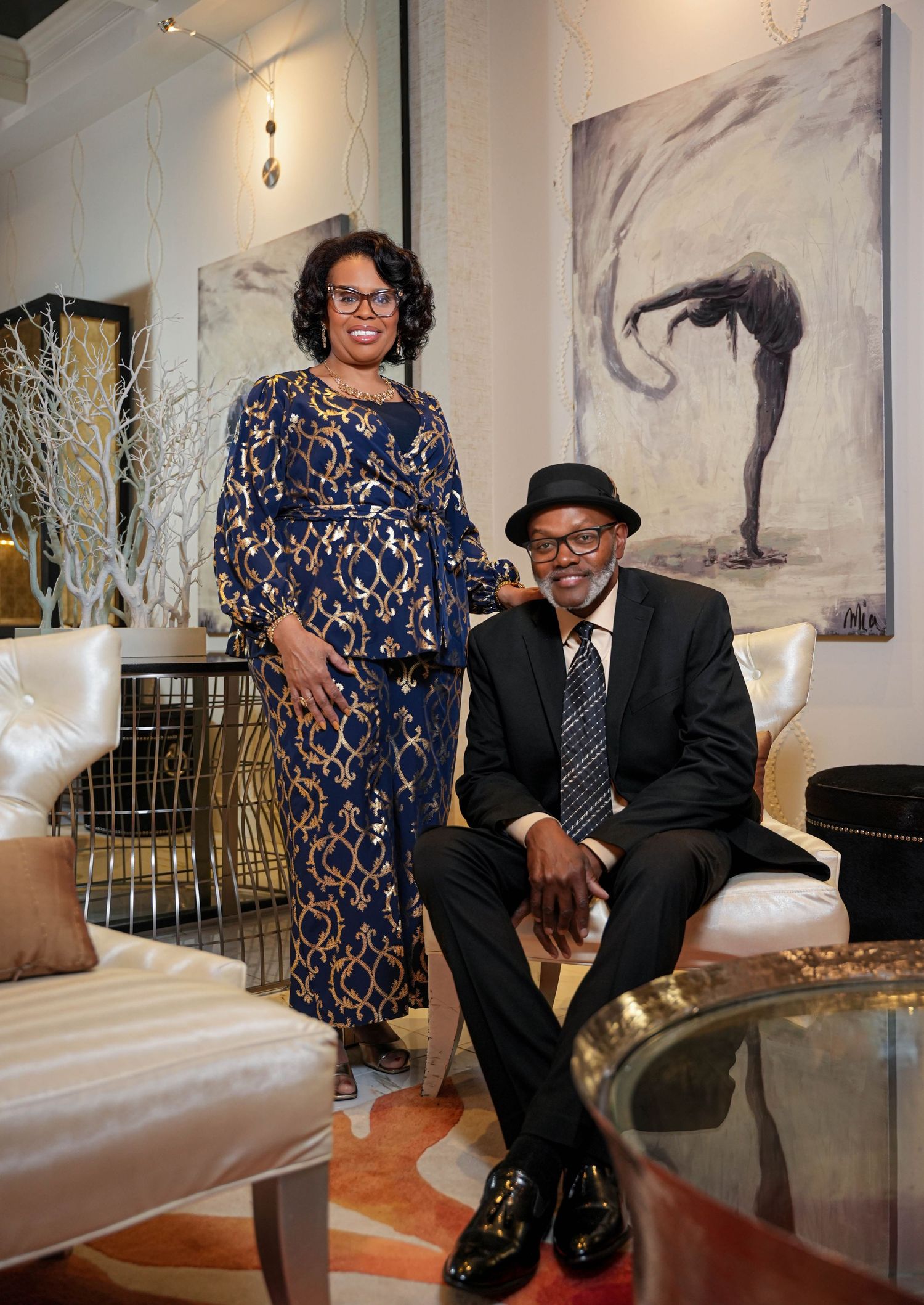 A well-dressed couple poses in an elegant living room with modern decor and artwork featuring a dancer on the wall.