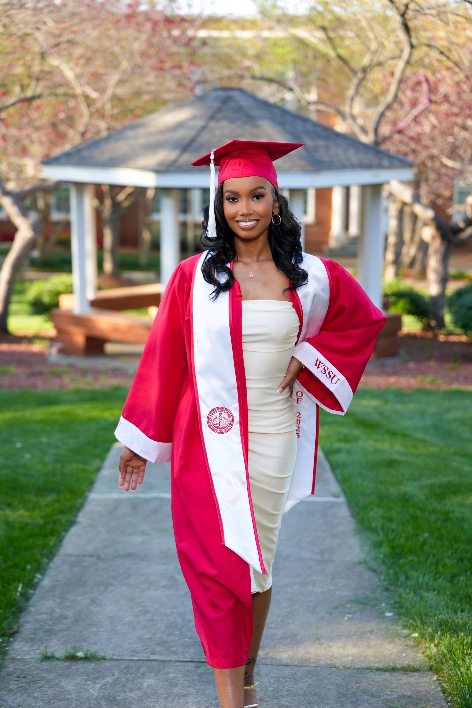 Graduate in red cap and gown walks along pathway with spring blossoms in background.