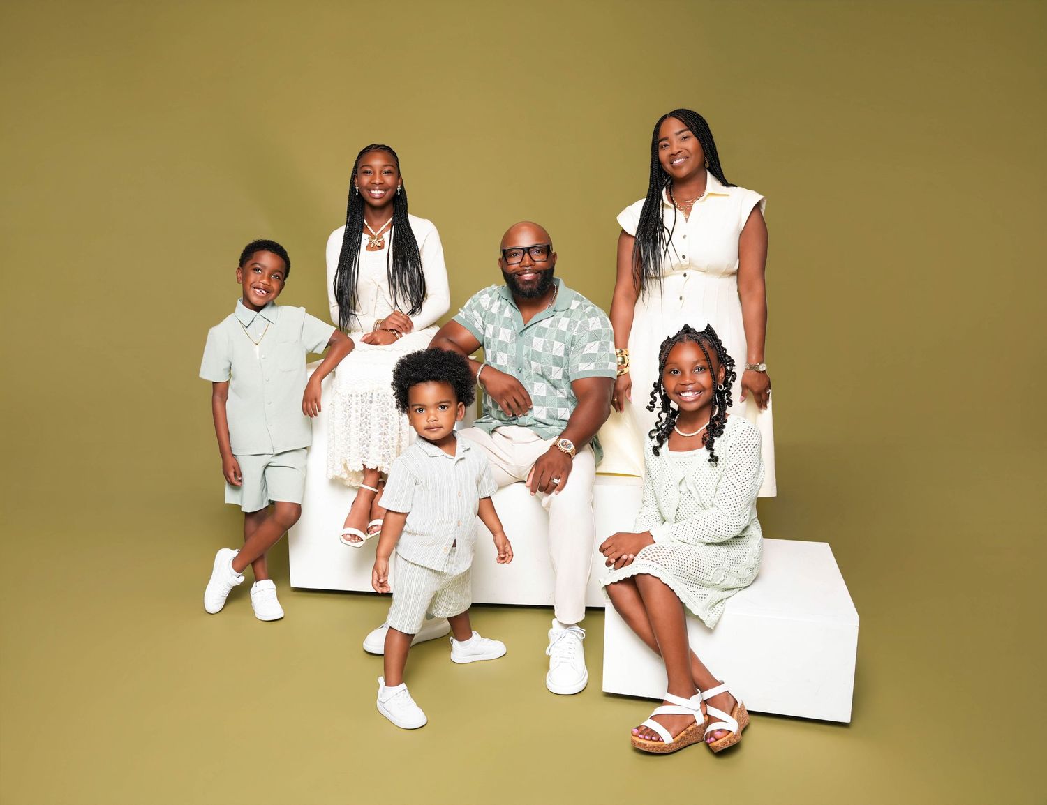 A joyful family of six poses together in coordinated white and mint outfits against a warm olive background.