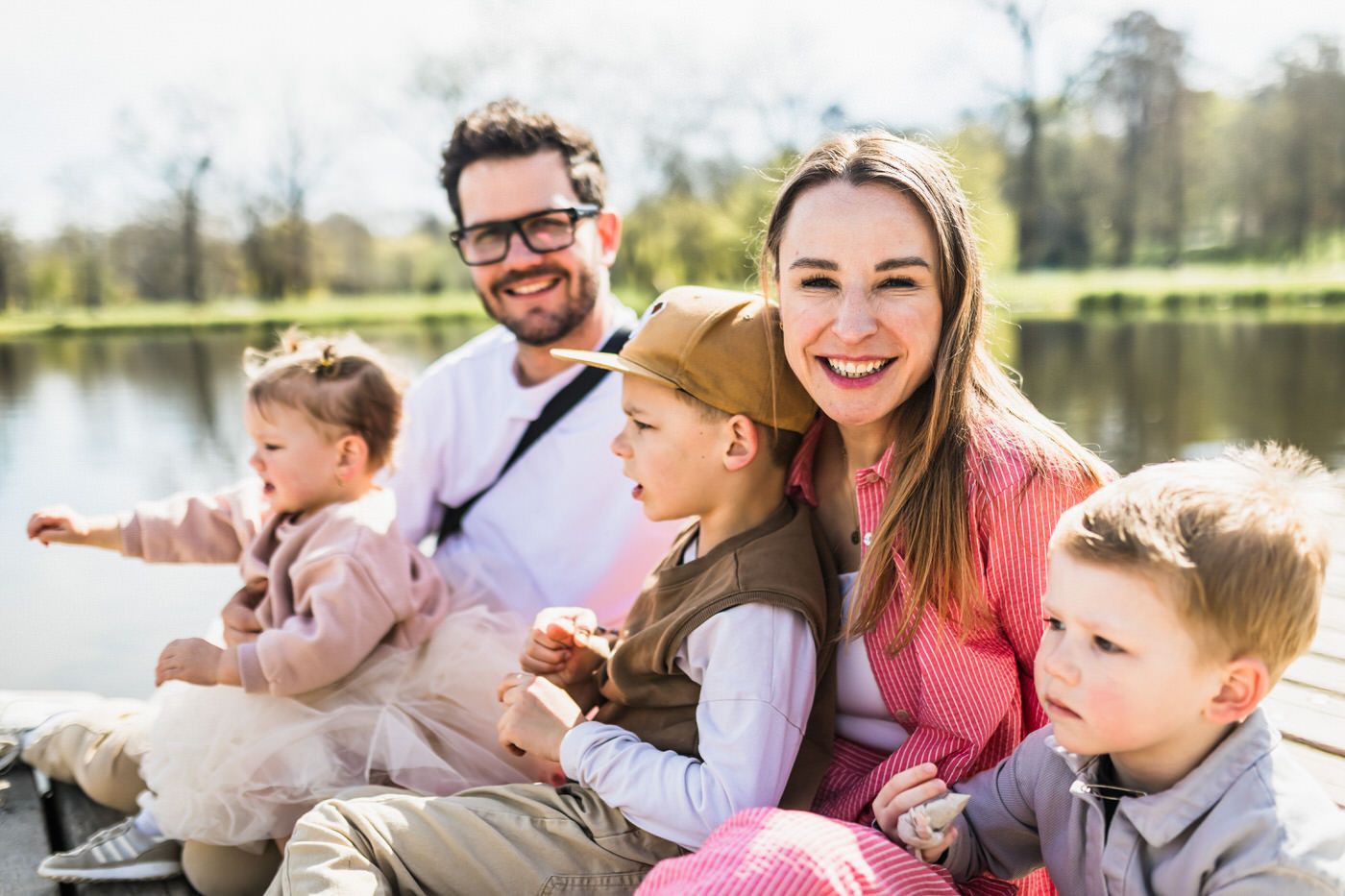 Happy family with three young children smiling outdoors by a sunny lake on a beautiful spring day.
