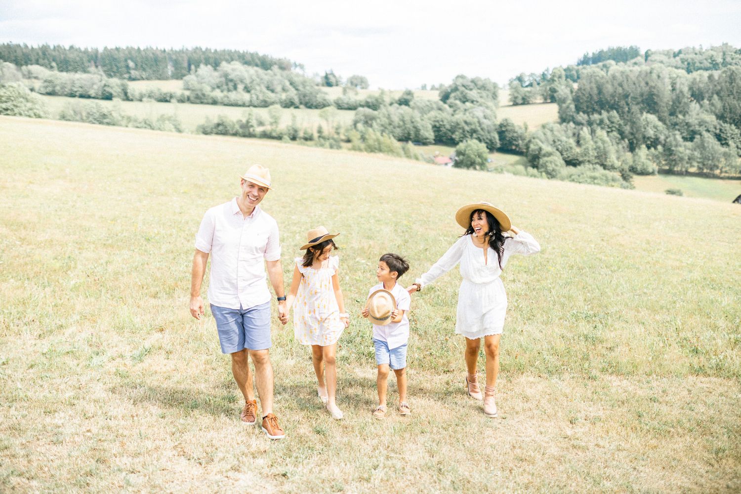 Family holding hands while walking through sunny meadow with forest in background.