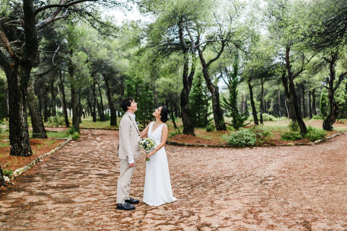 A couple shares a romantic moment on a winding dirt path surrounded by lush green trees in a forest setting.