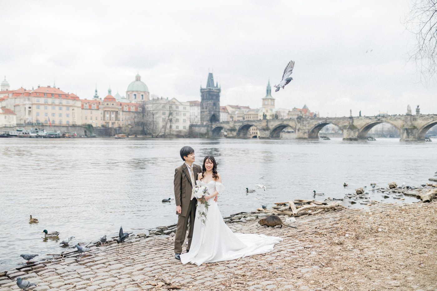 A romantic couple poses for wedding photos along the scenic riverbank of Prague's Charles Bridge on a cloudy day.