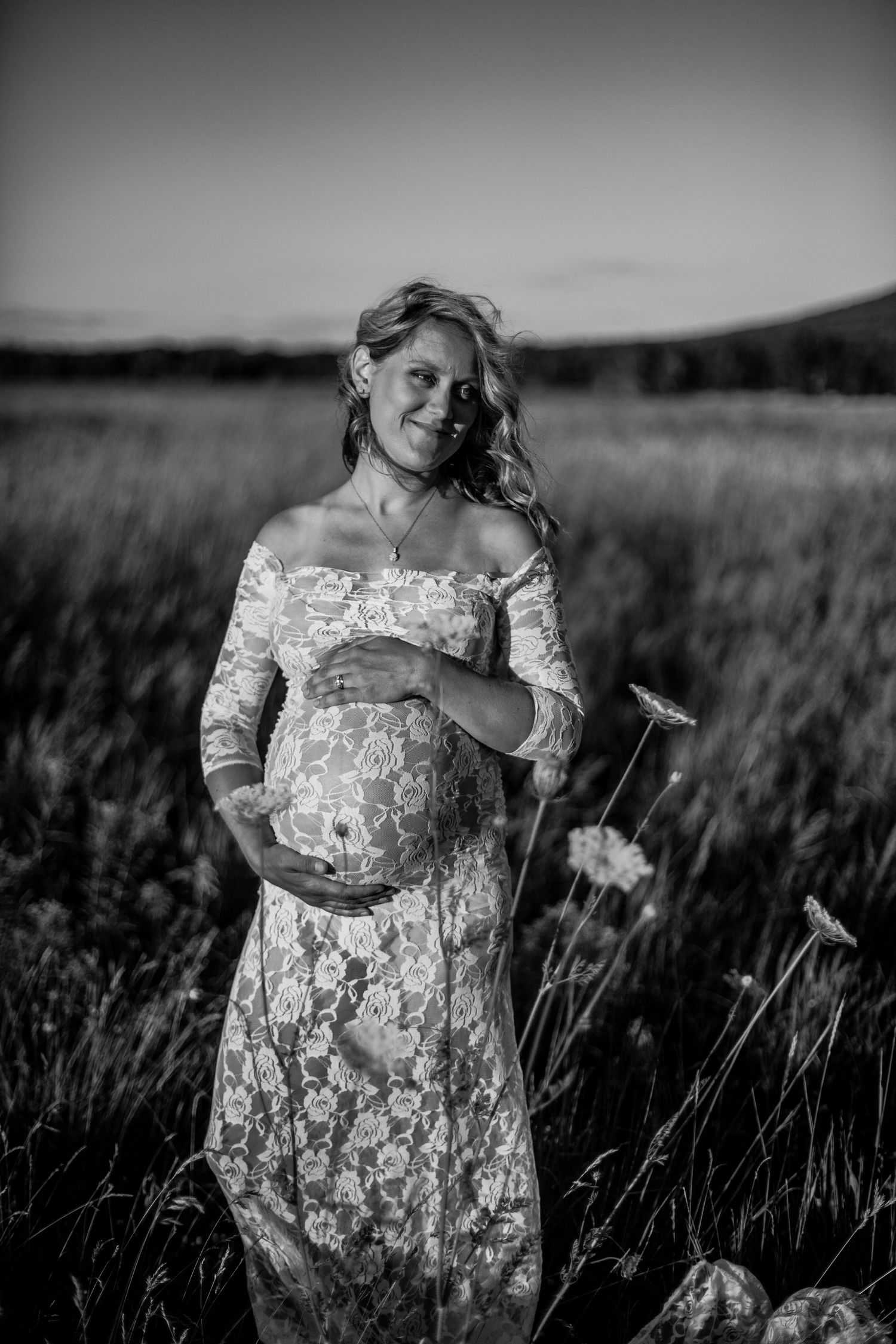 Smiling pregnant woman in lace dress cradling baby bump in a field at dusk, black and white maternity photo.