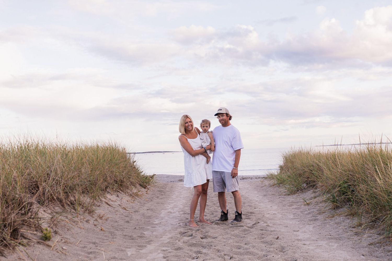 Happy family of three posing on a sandy Cape Cod beach path surrounded by tall grass.