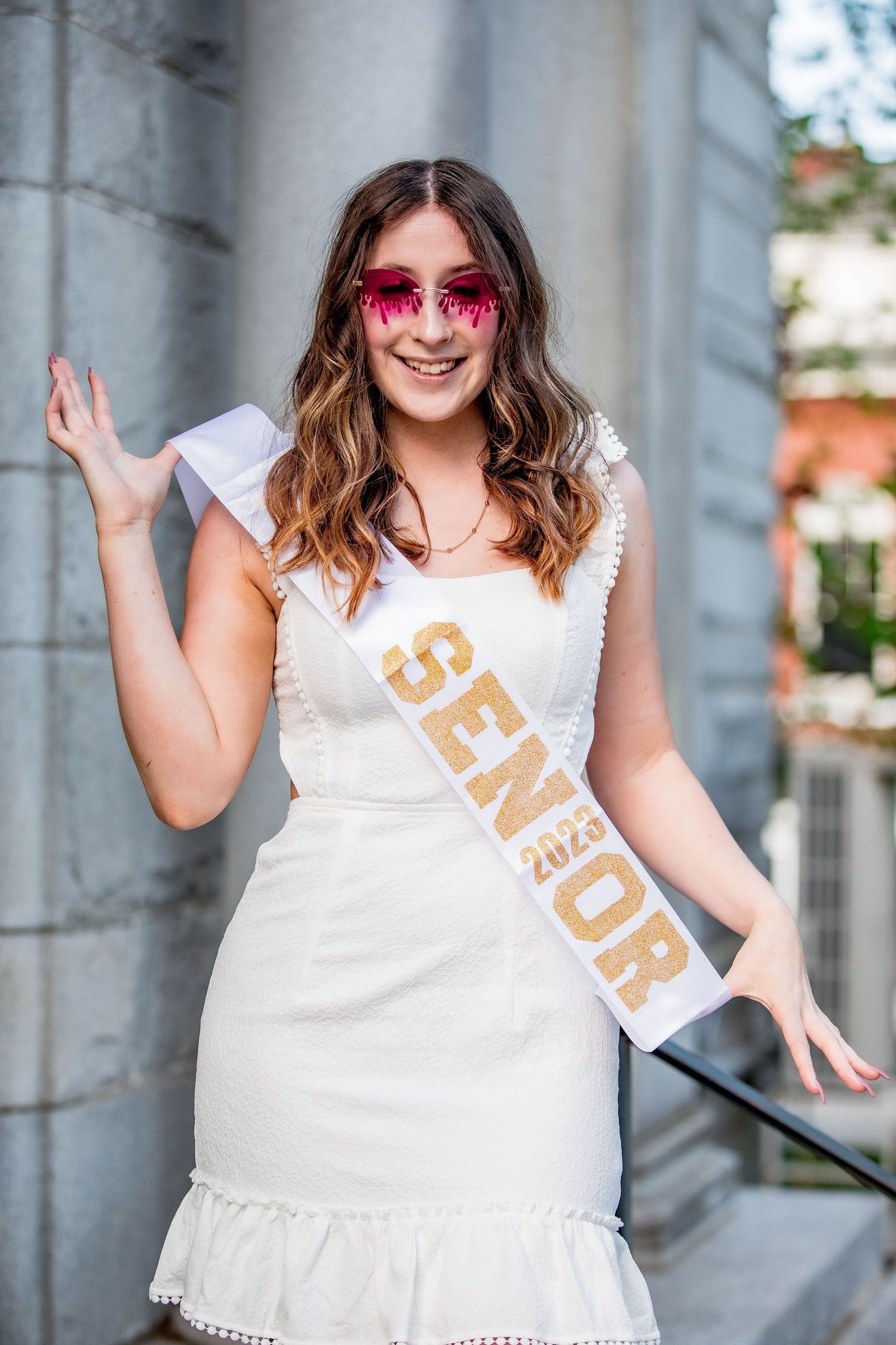 Smiling young woman in white dress wearing a Senior 2023 sash and pink sunglasses at graduation.
