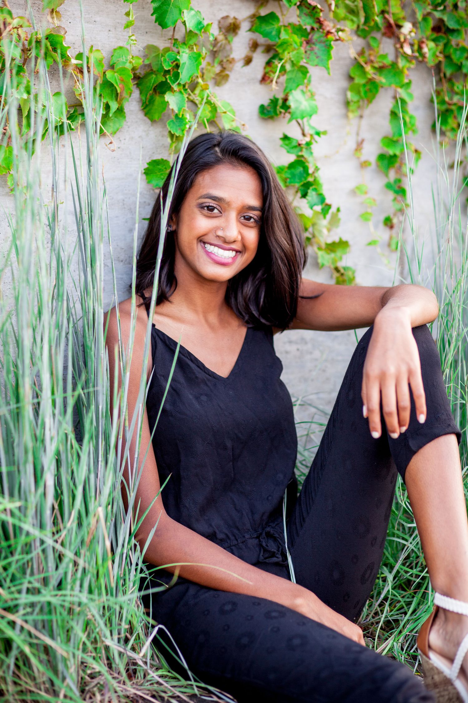 Smiling South Asian woman in black outfit sits among tall grass and ivy against concrete wall.