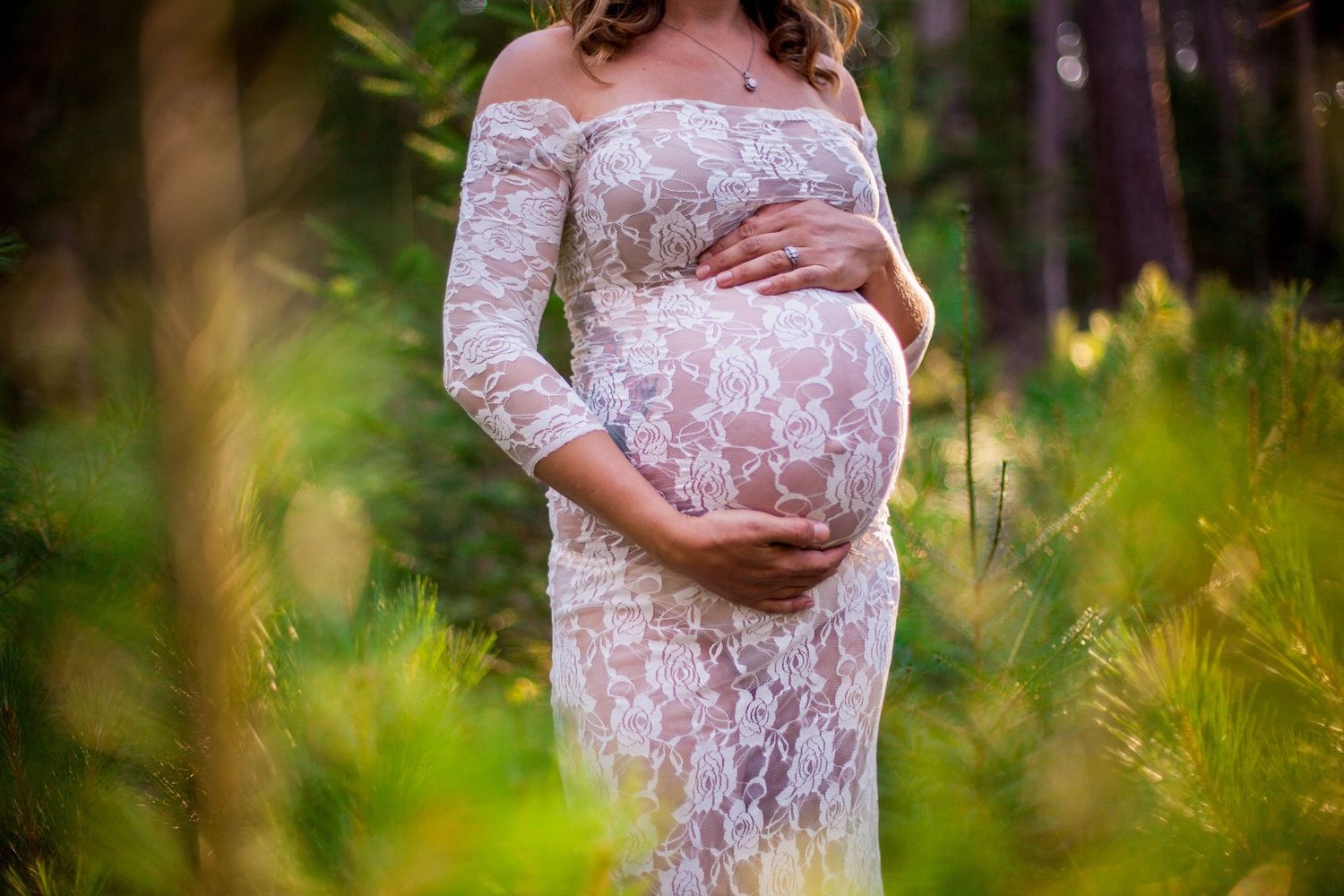 Pregnant woman in white lace gown cradling baby bump outdoors in green forest.