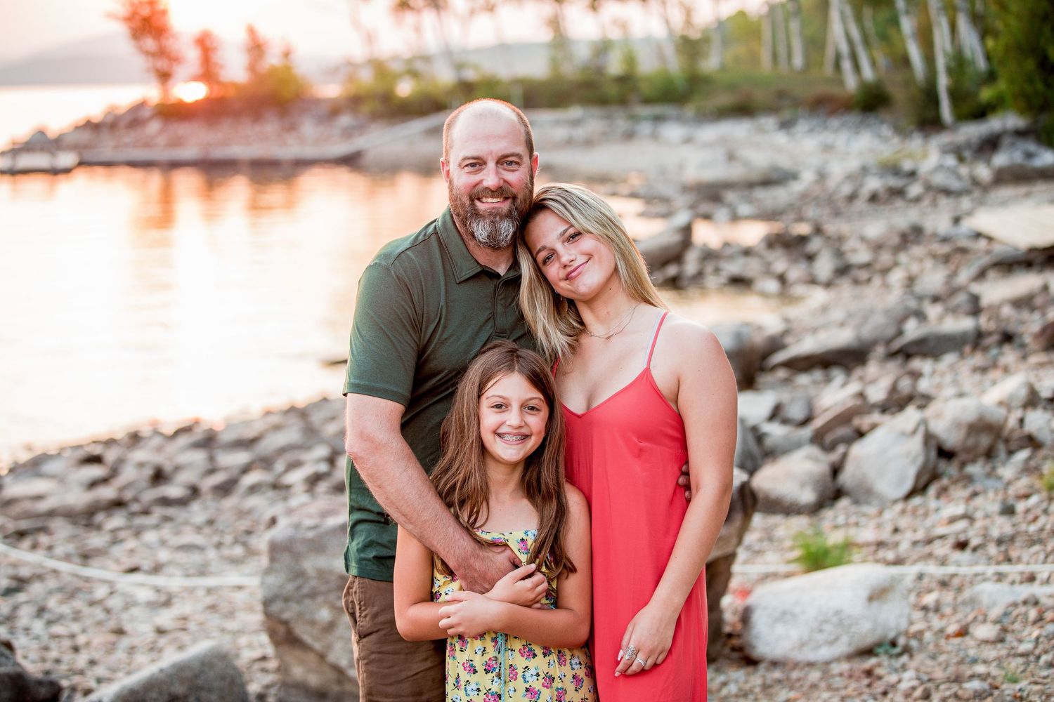 Family of three smiling at sunset on a rocky lakeside shore.
