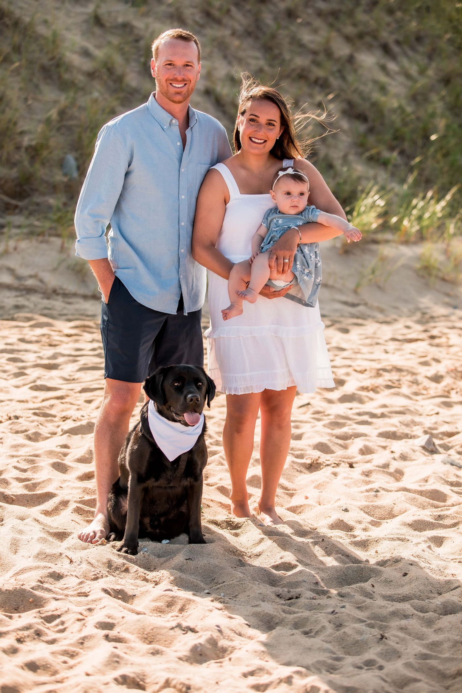 Smiling couple with baby and black Labrador dog posing on sandy Cape Cod beach at sunset.