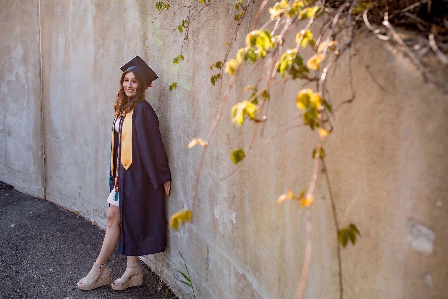 Young woman in navy graduation cap and gown leaning against a concrete wall with autumn foliage.