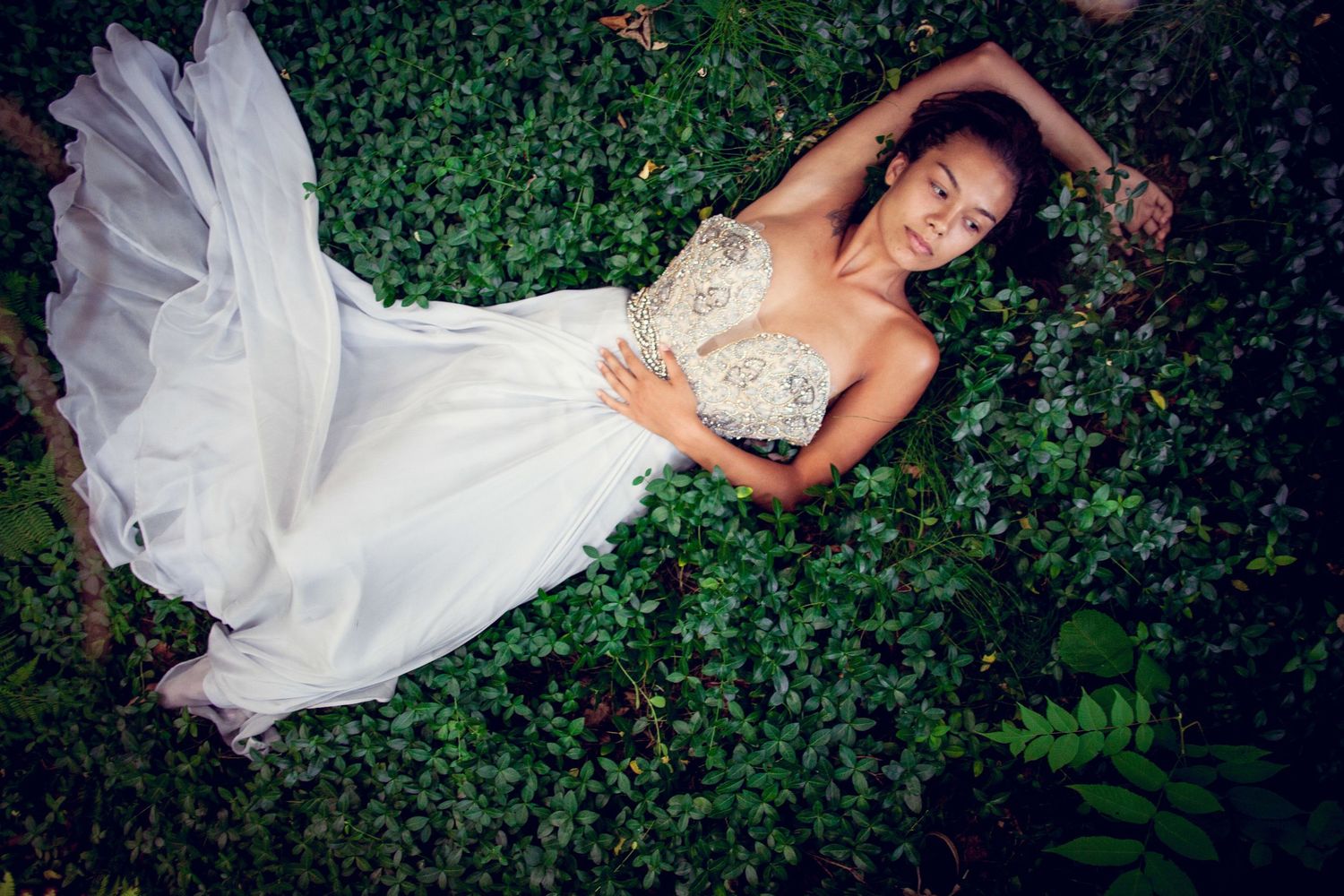 Woman in elegant white beaded bridal gown lying gracefully on lush green garden foliage.