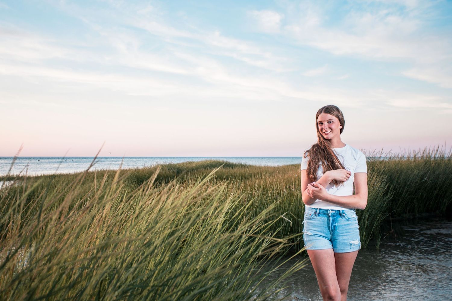 Young woman in white tee and denim shorts smiling on sandy beach path surrounded by tall grass at sunset.